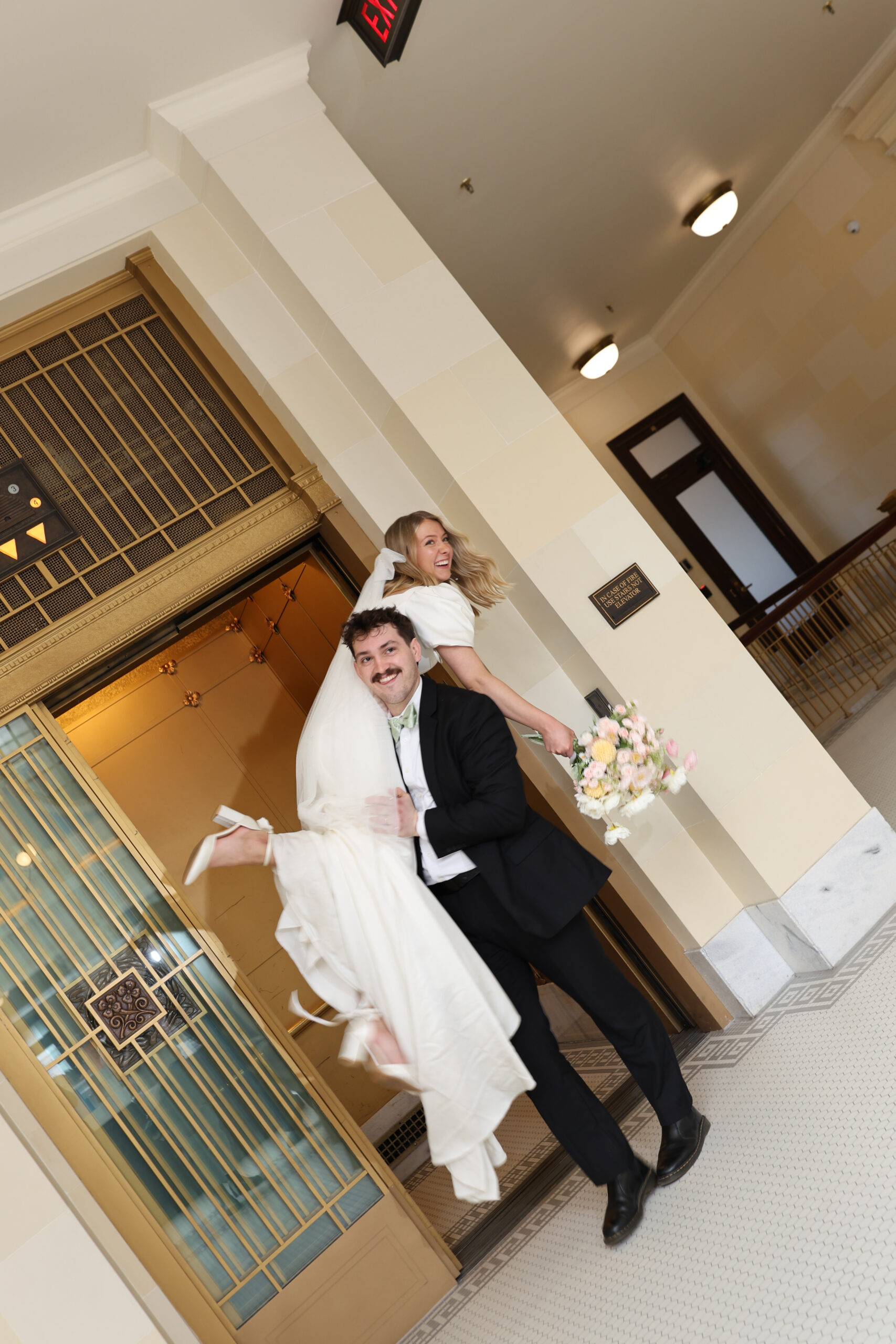 A playful moment as a groom carries his bride through ornate golden elevator doors, photographed by a travel elopement photographer.