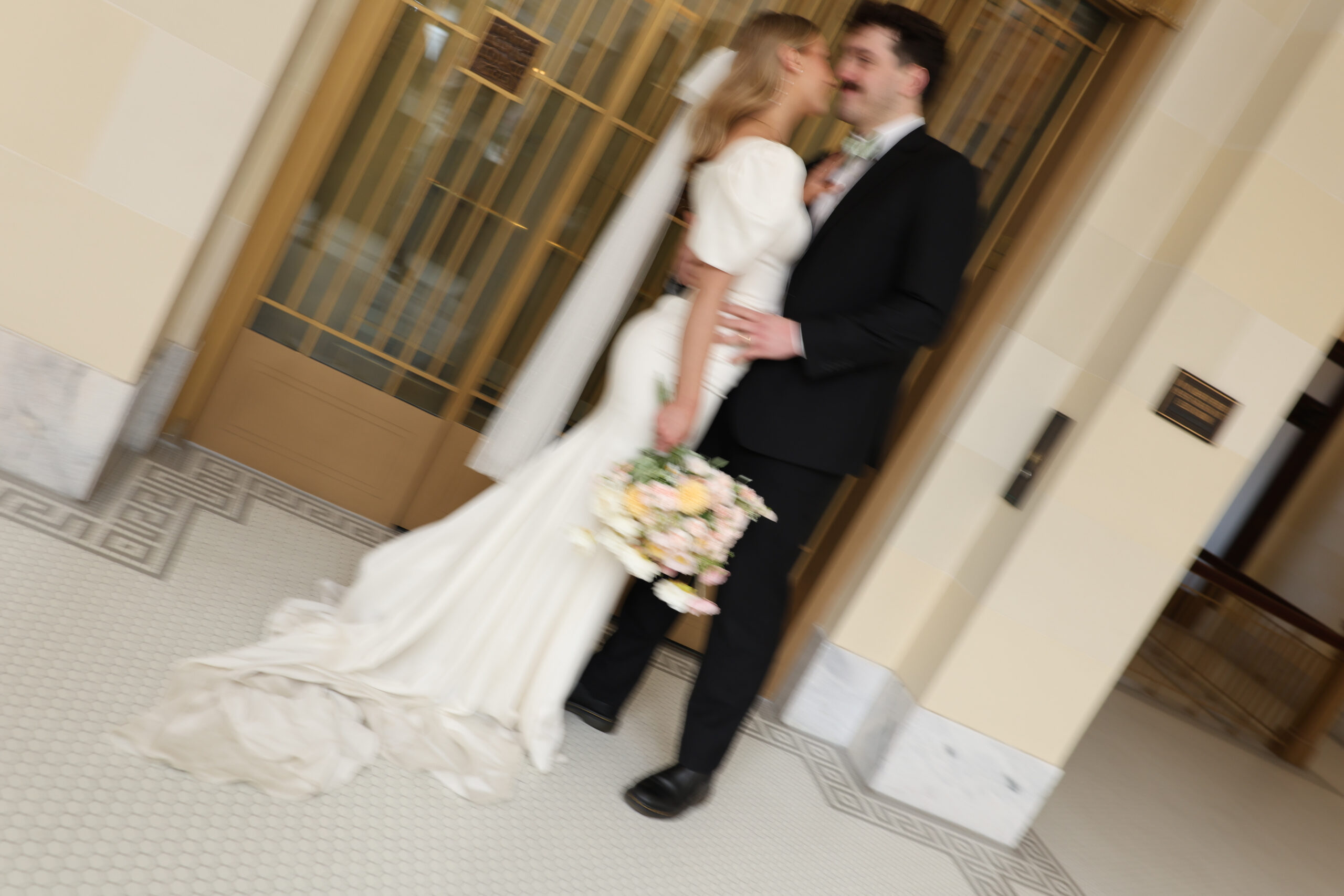 Blurry shot of a bride holding her bouquet as she kisses her groom in front of vintage gold elevator doors.