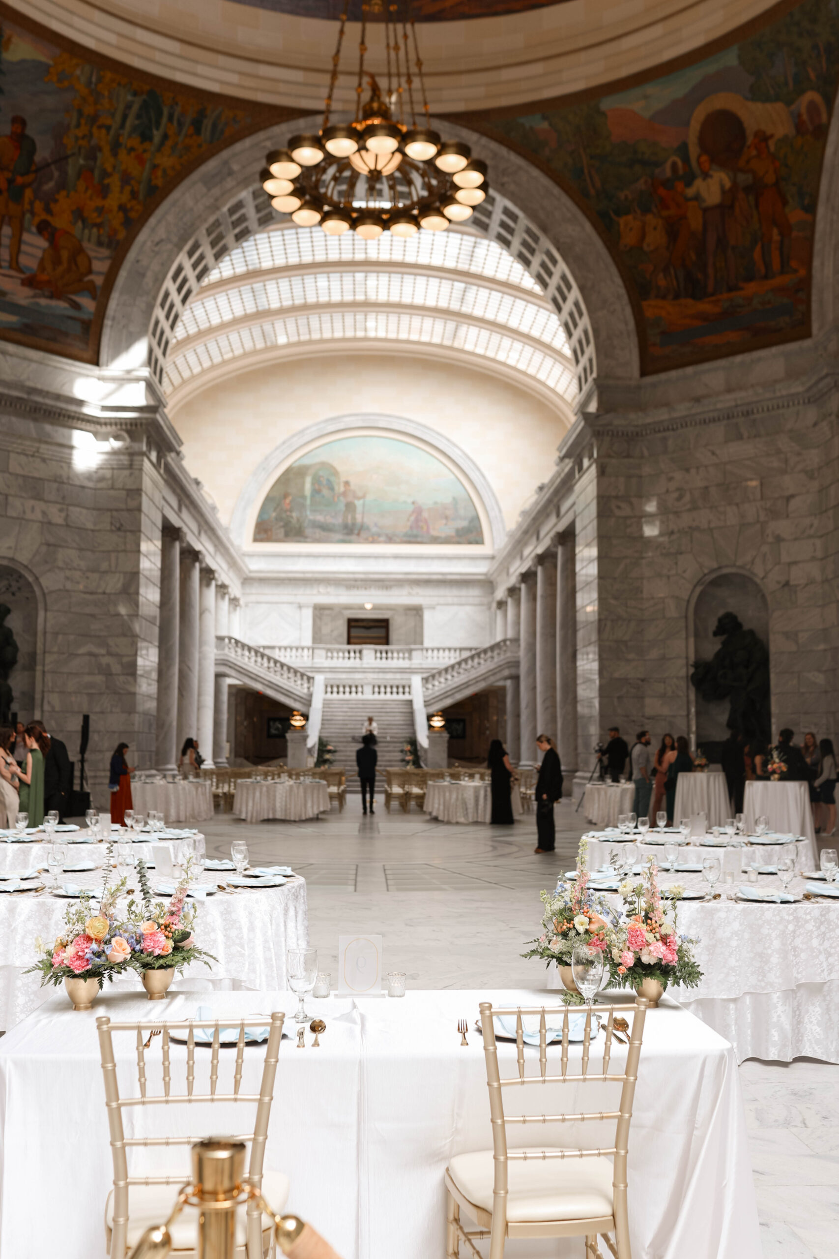 Elegant wedding reception setup inside a historic building, with round tables, soft floral centerpieces, and gold chairs under ornate chandeliers.