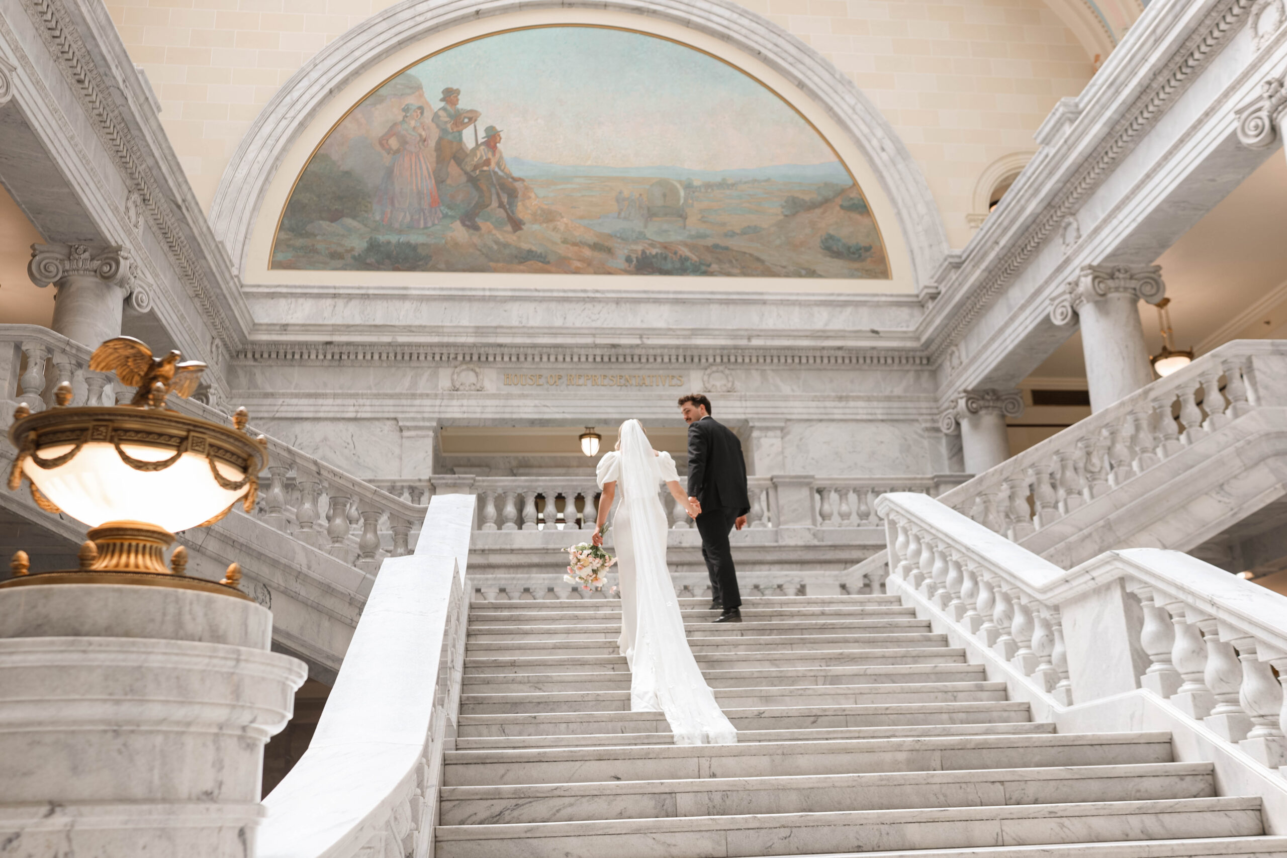 Couple holding hands while walking up grand marble stairs with ornate murals above them.