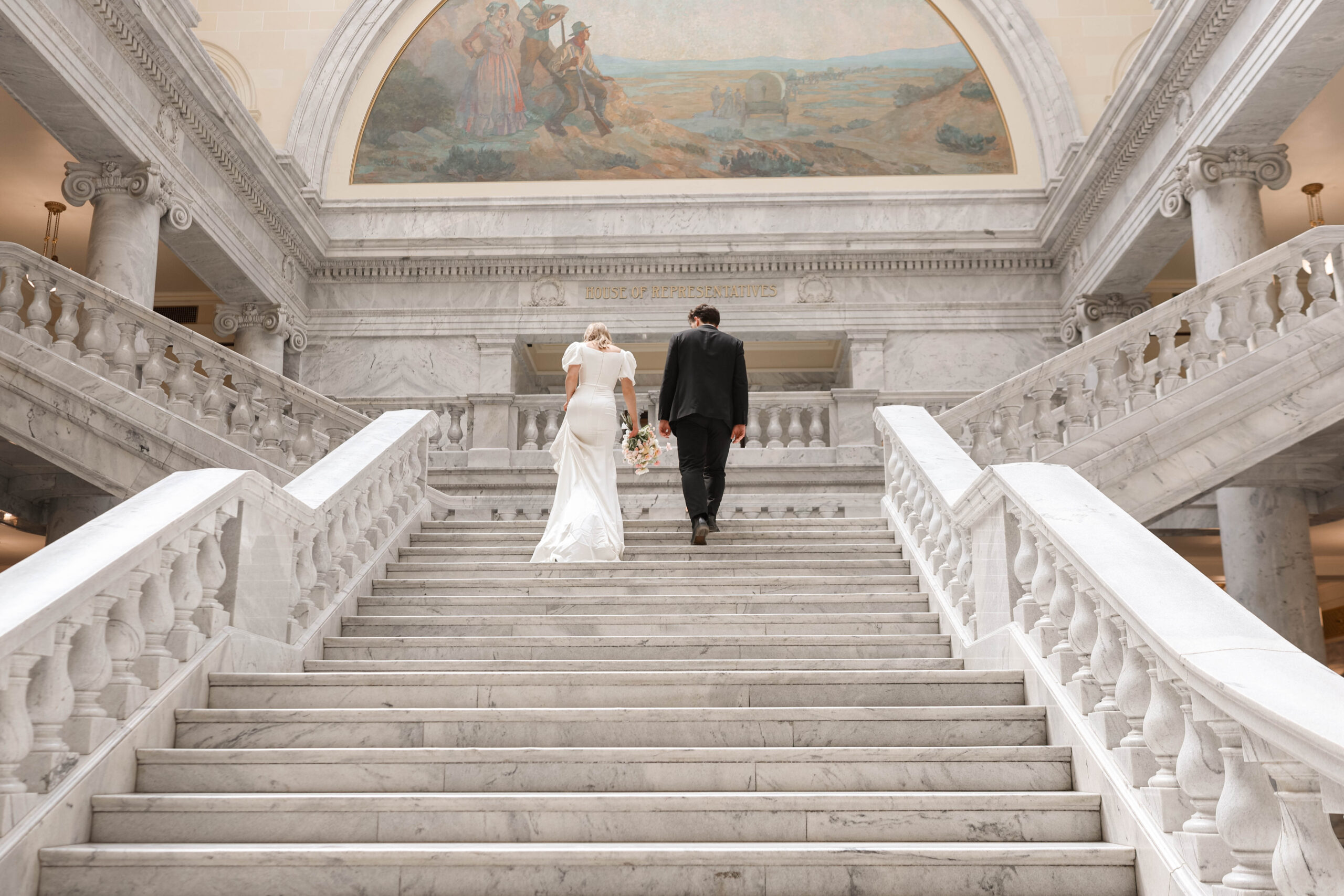 Bride and groom walk hand-in-hand up grand marble stairs inside the Utah Capitol building, their modern elopement unfolding in a stunning architectural space.