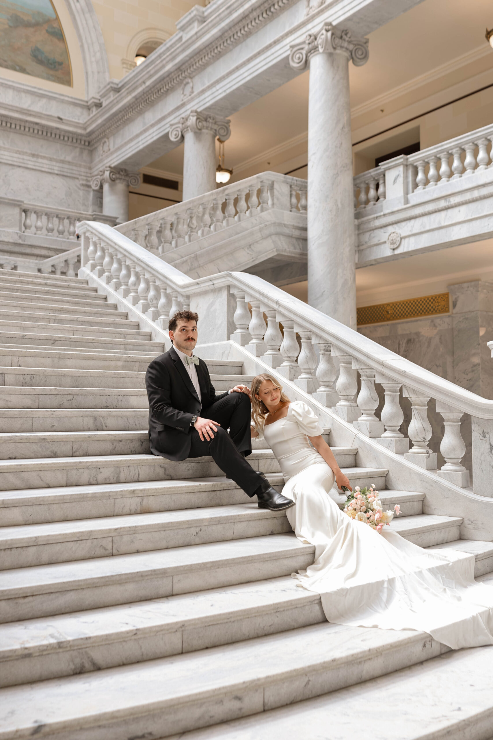 Bride and groom share a relaxed moment sitting together on marble stairs, her bouquet resting beside them.