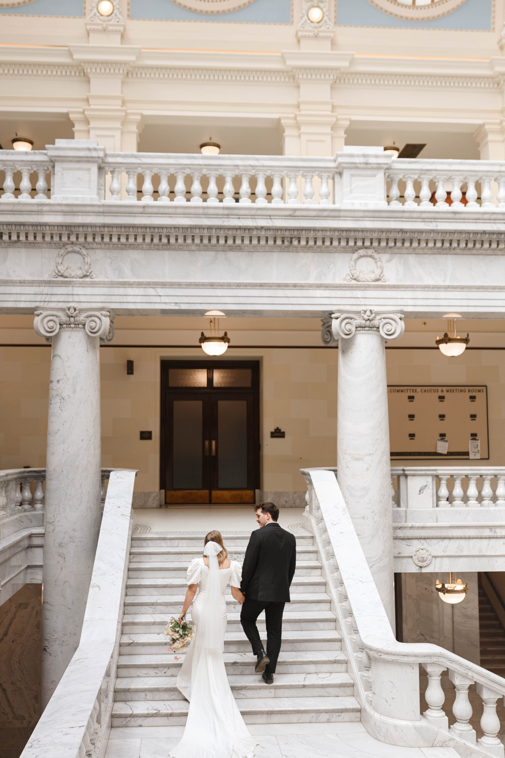 Romantic moment of newlyweds walking hand-in-hand up marble stairs, bouquet in hand.