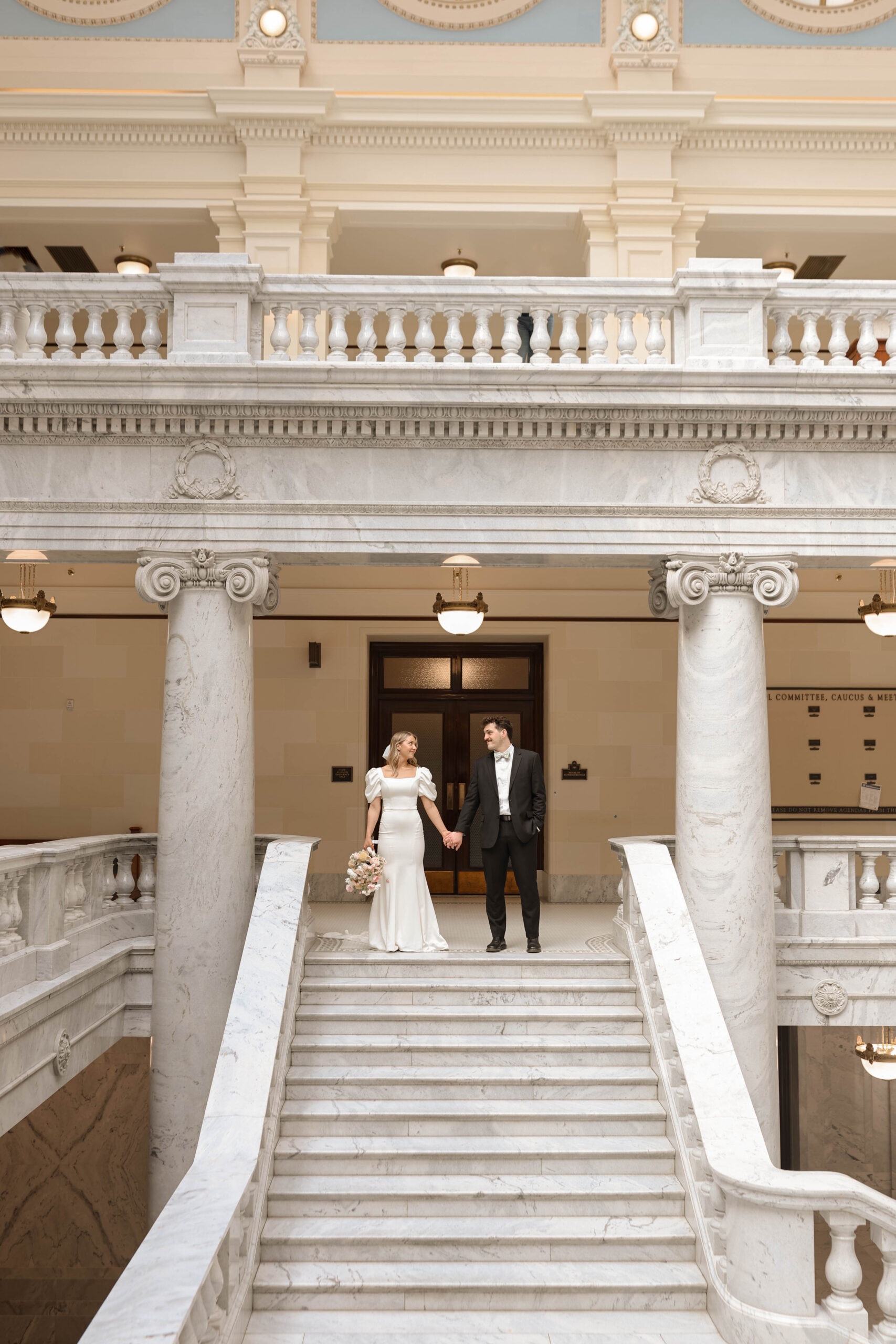 Bride and groom hold hands on a balcony inside the Utah Capitol, framed by classic marble columns and ornate architecture.