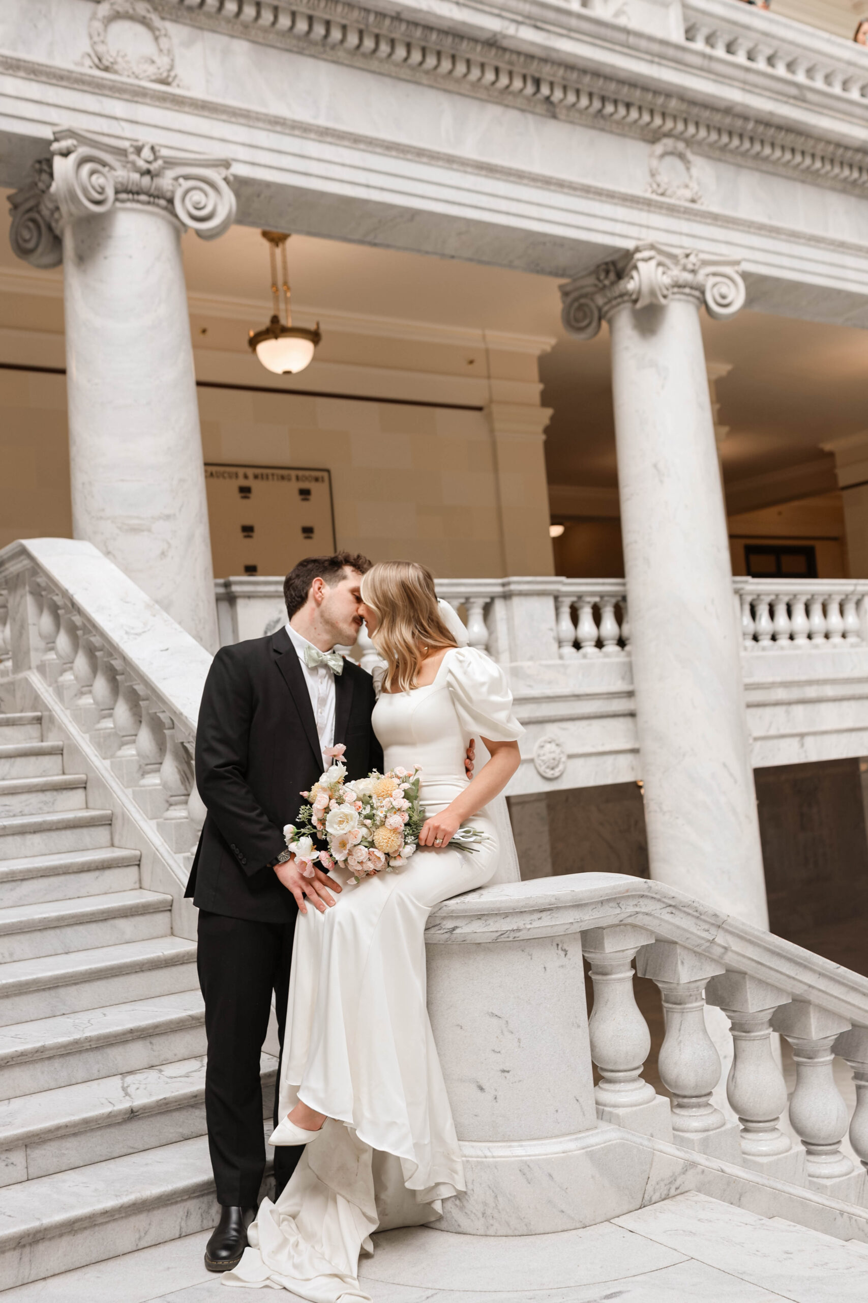 Bride and groom sit on a marble railing, sharing a quiet kiss after their ceremony, bouquet in hand.