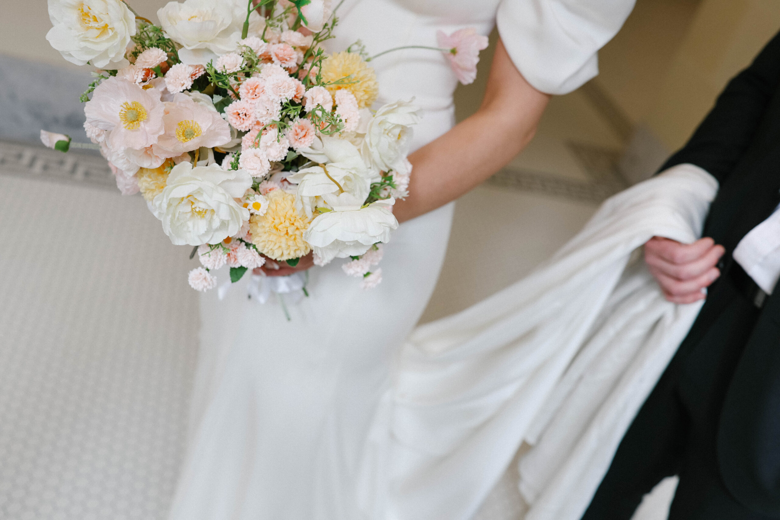 Close-up of a modern bridal bouquet featuring soft yellow and blush flowers, held by the bride in a sleek white gown.