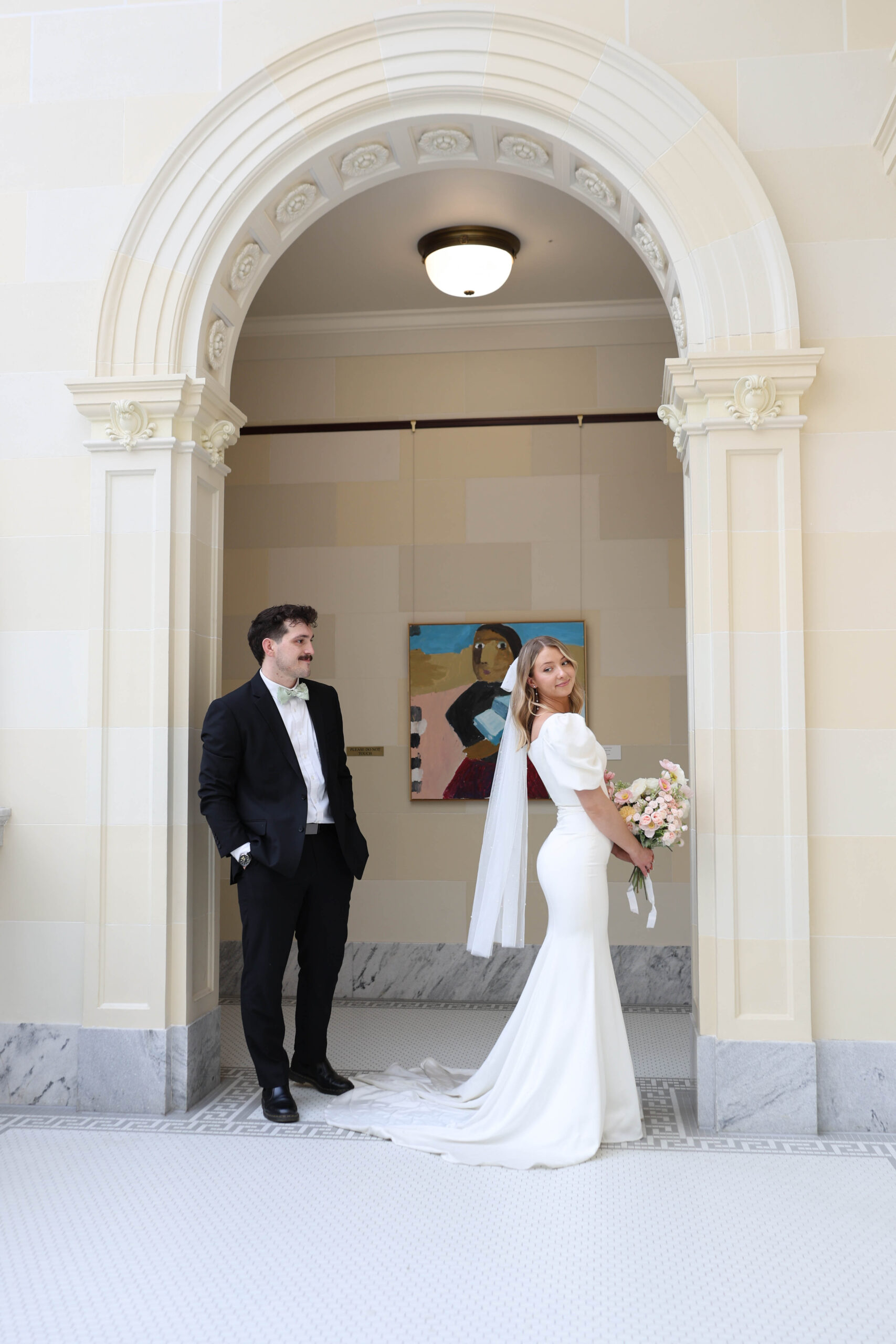 Bride holding her bouquet while glancing over her shoulder at her groom under a marble archway, captured by a travel elopement photographer.