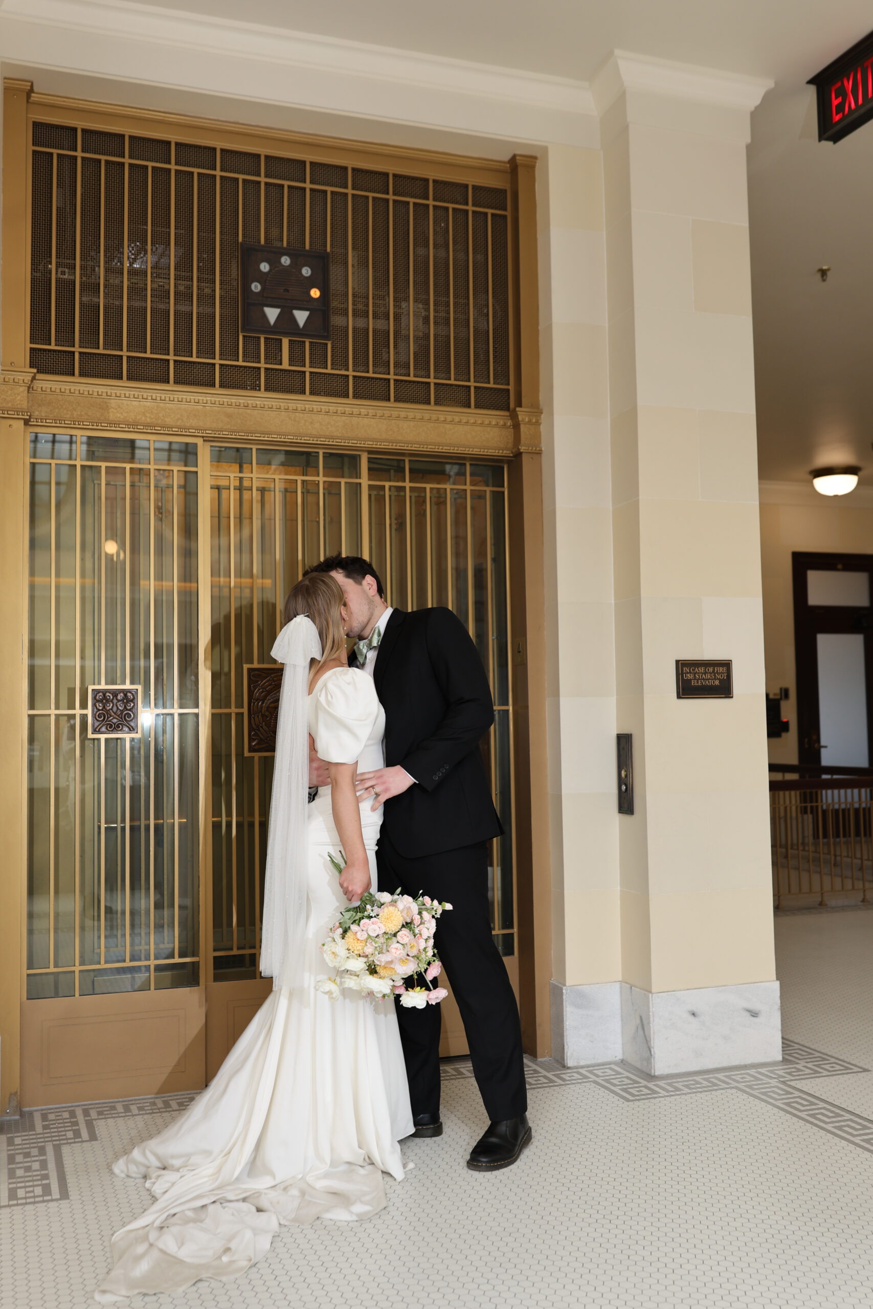 Bride and groom sharing a kiss in front of golden elevator doors, flowers held between them.