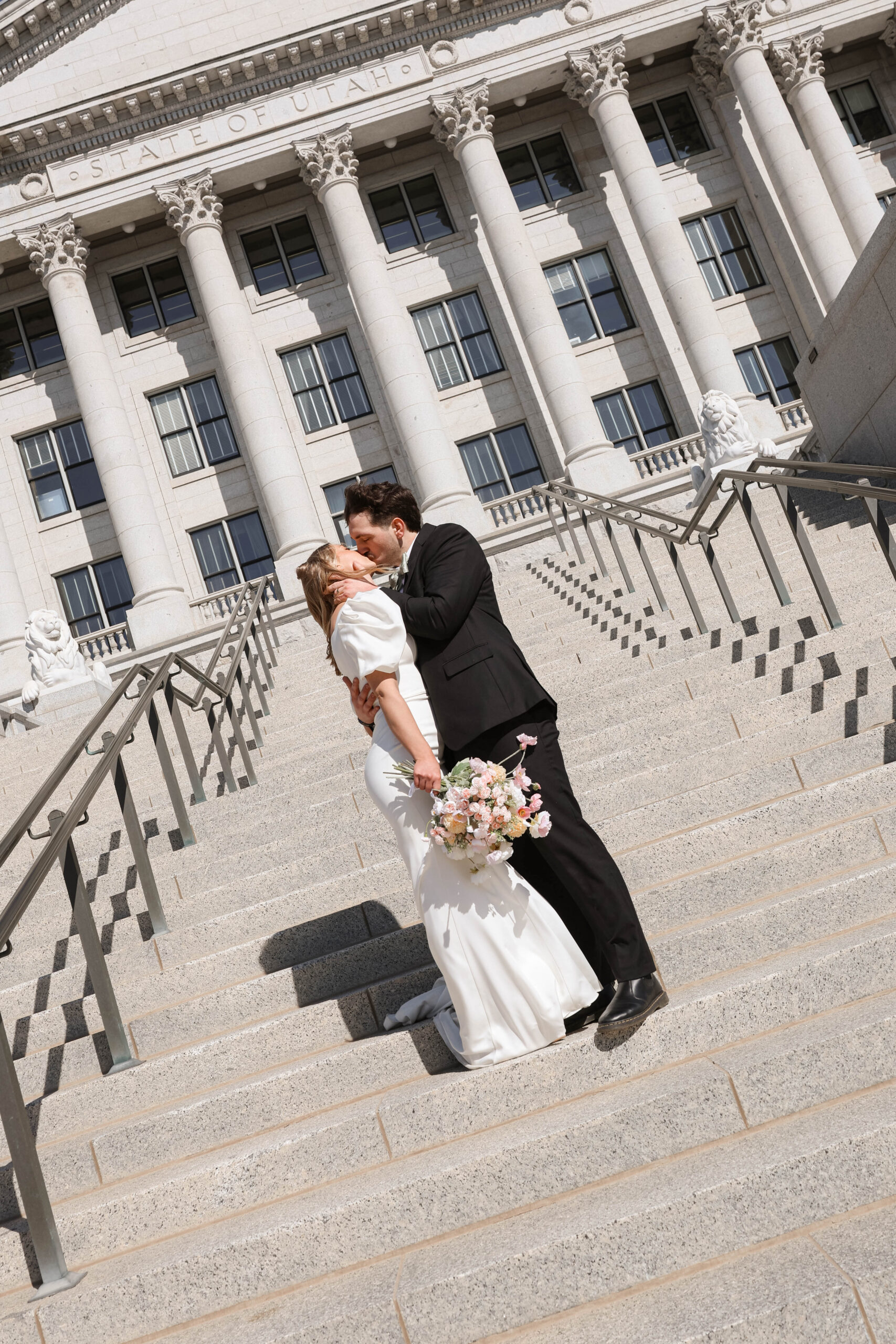 A travel elopement photographer documents the bride and groom sharing a kiss on the stone steps outside the Utah State Capitol, bouquet in hand.