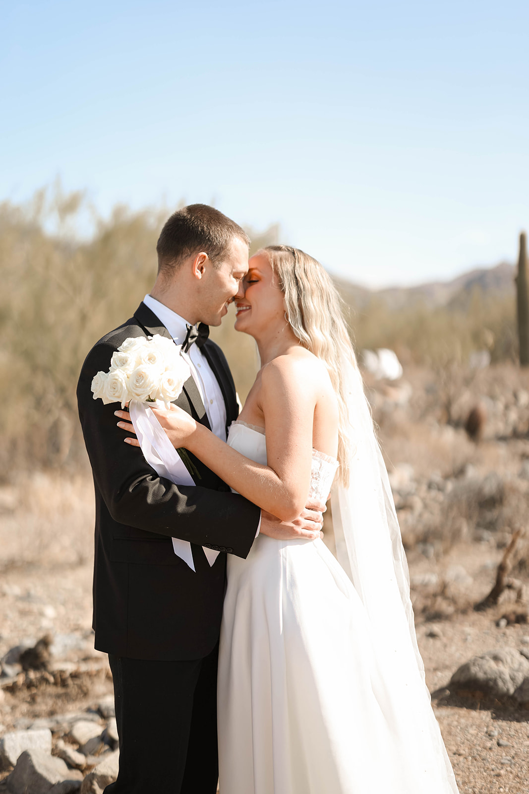 A bride and groom sharing a forehead-to-forehead moment while holding a white rose bouquet in the middle of a sun-drenched desert landscape.