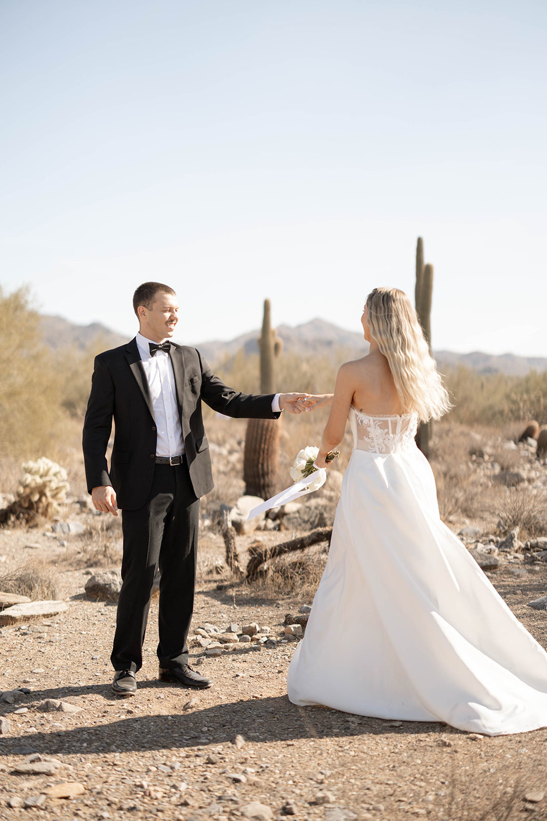 Groom reaches out to his bride as they stand among towering cacti, a romantic moment captured in one of the most scenic Arizona elopement locations.