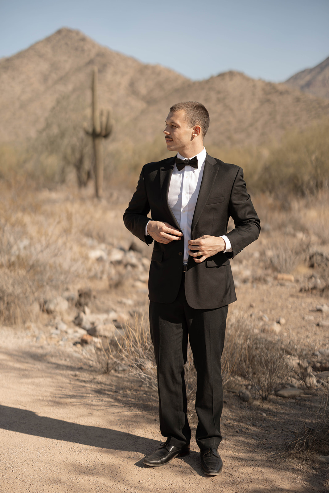 Groom adjusting his jacket while standing among cacti and desert mountains, showcasing the classic style fit for Arizona elopement locations.