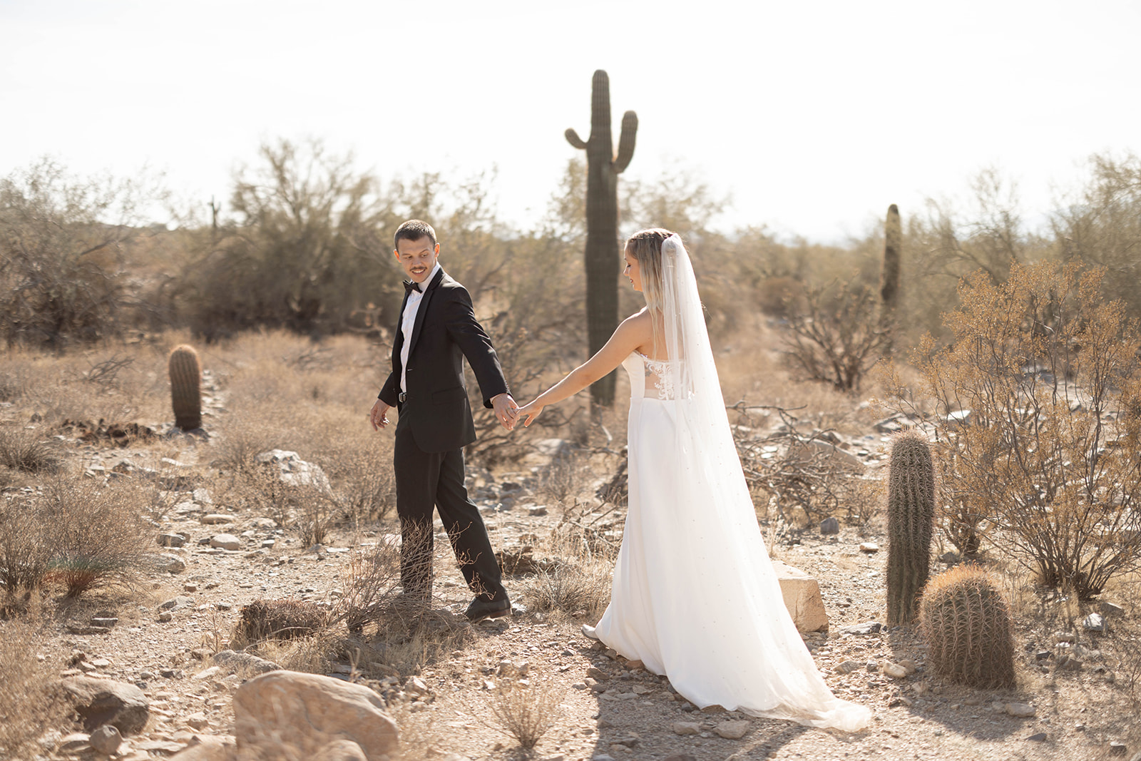 Groom holding the bride’s hand, leading her through a rocky desert landscape filled with saguaro cacti.