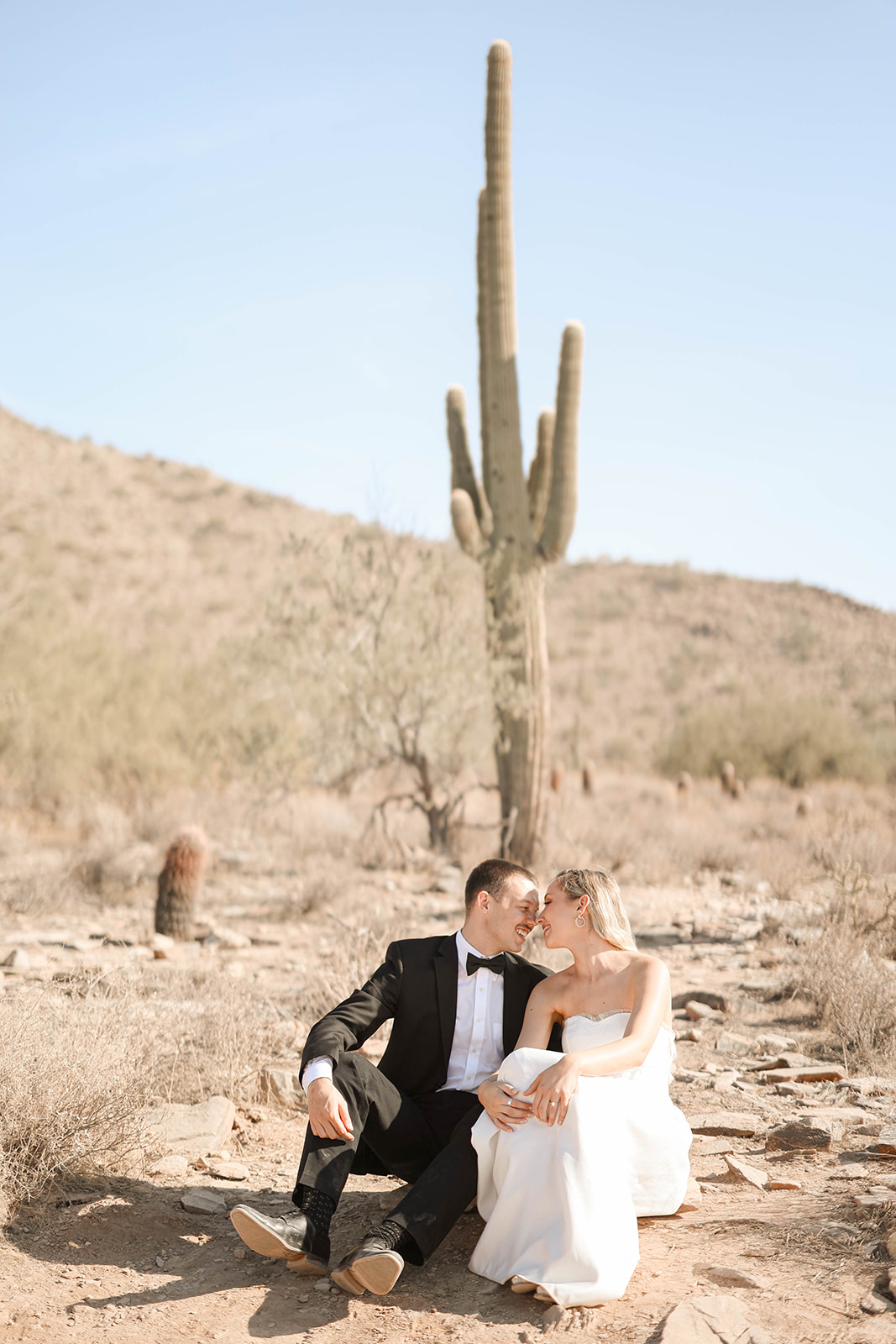 Bride and groom sit close together in the Arizona desert, sharing an intimate moment with a tall saguaro cactus behind them. The groom wears a black tuxedo and the bride is in a strapless white gown, both looking relaxed and joyful in the natural setting.
