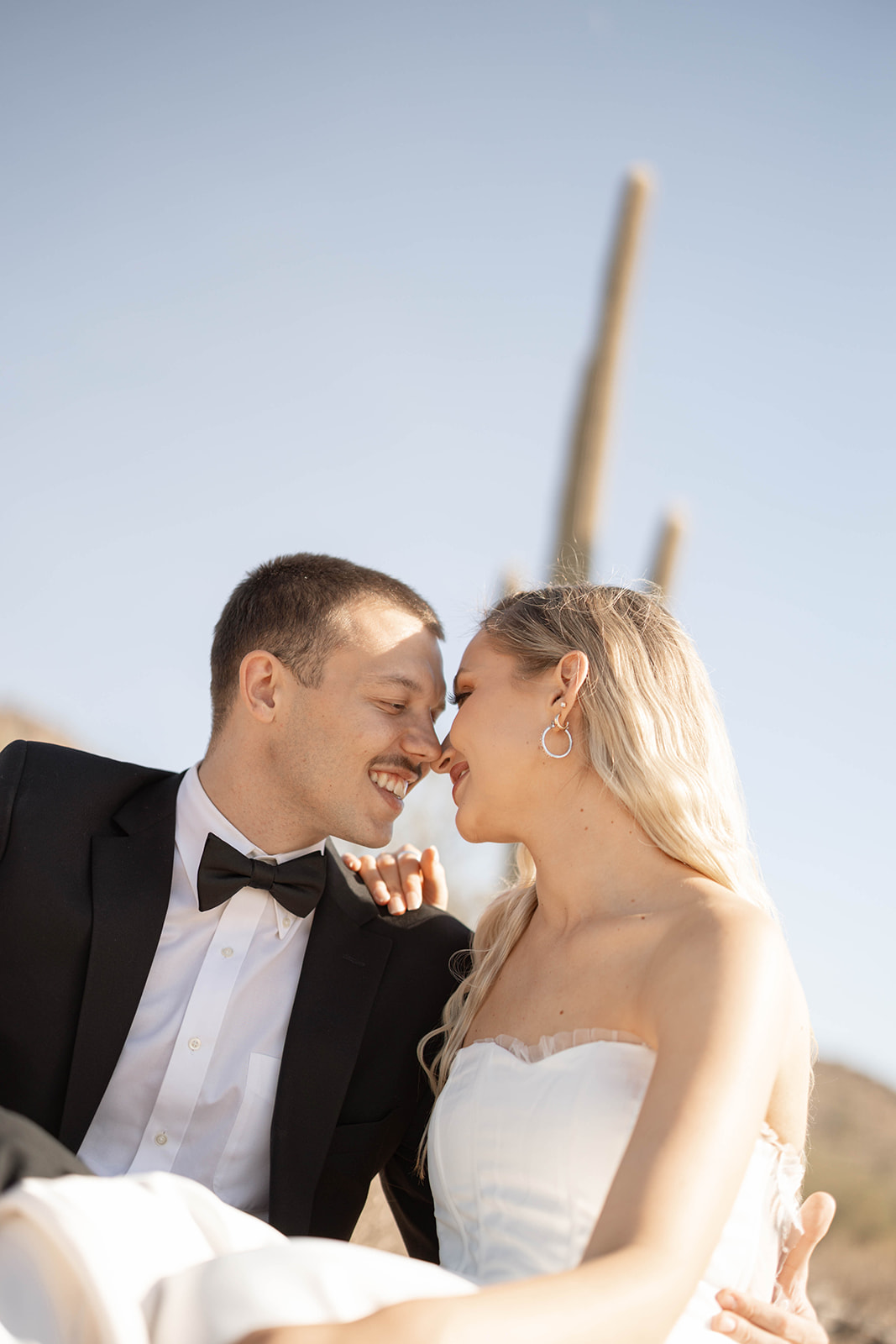 Playful moment as the bride and groom lean in, nose-to-nose, smiling beneath a clear desert sky.