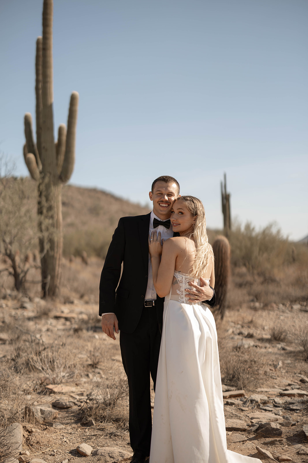 Standing together with arms wrapped around each other, the newlyweds smile against a backdrop of mountains and desert terrain, a timeless Arizona elopement location.