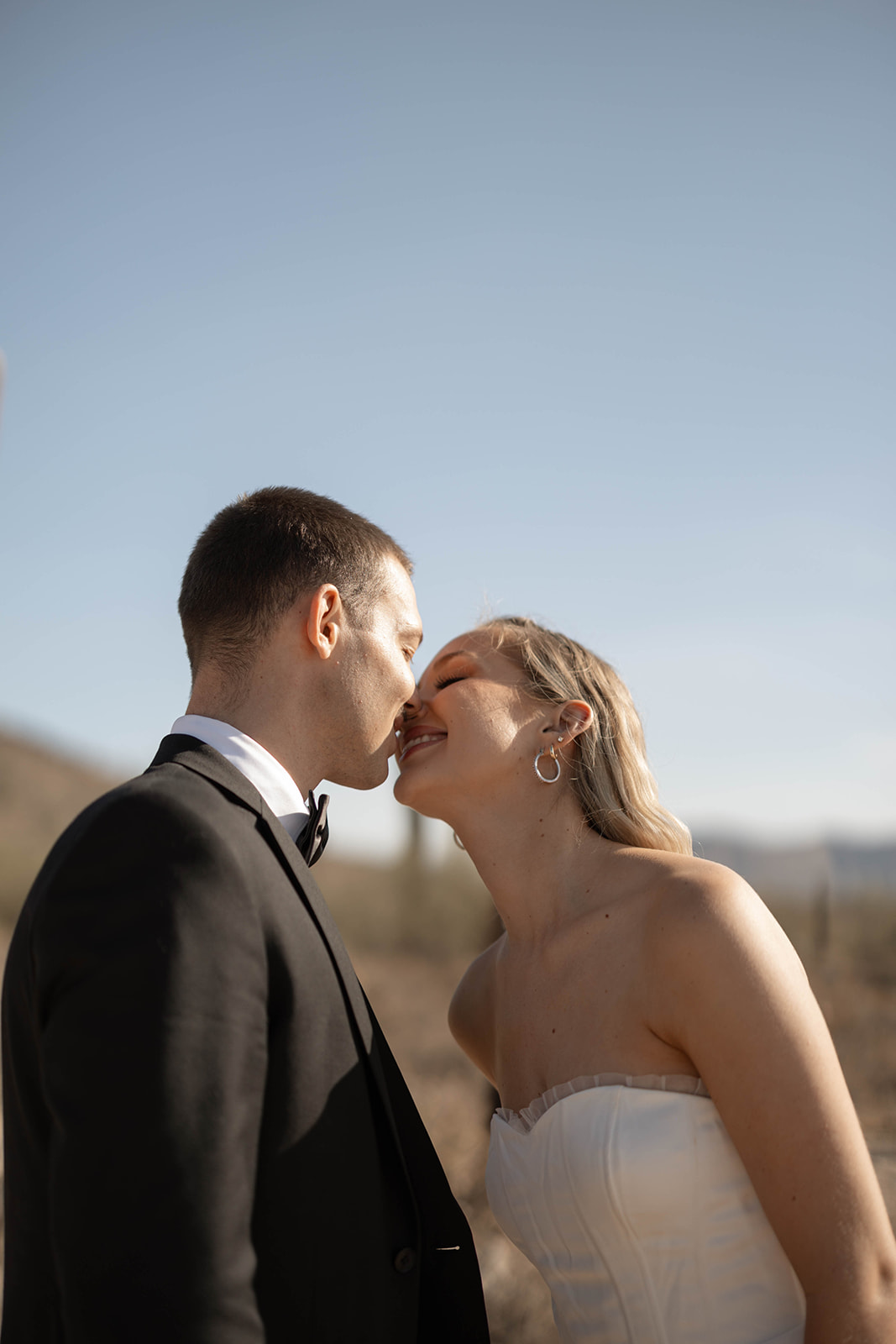 A bride and groom share a joyful almost-kiss in the Arizona desert, sunlit and surrounded by wide-open skies.