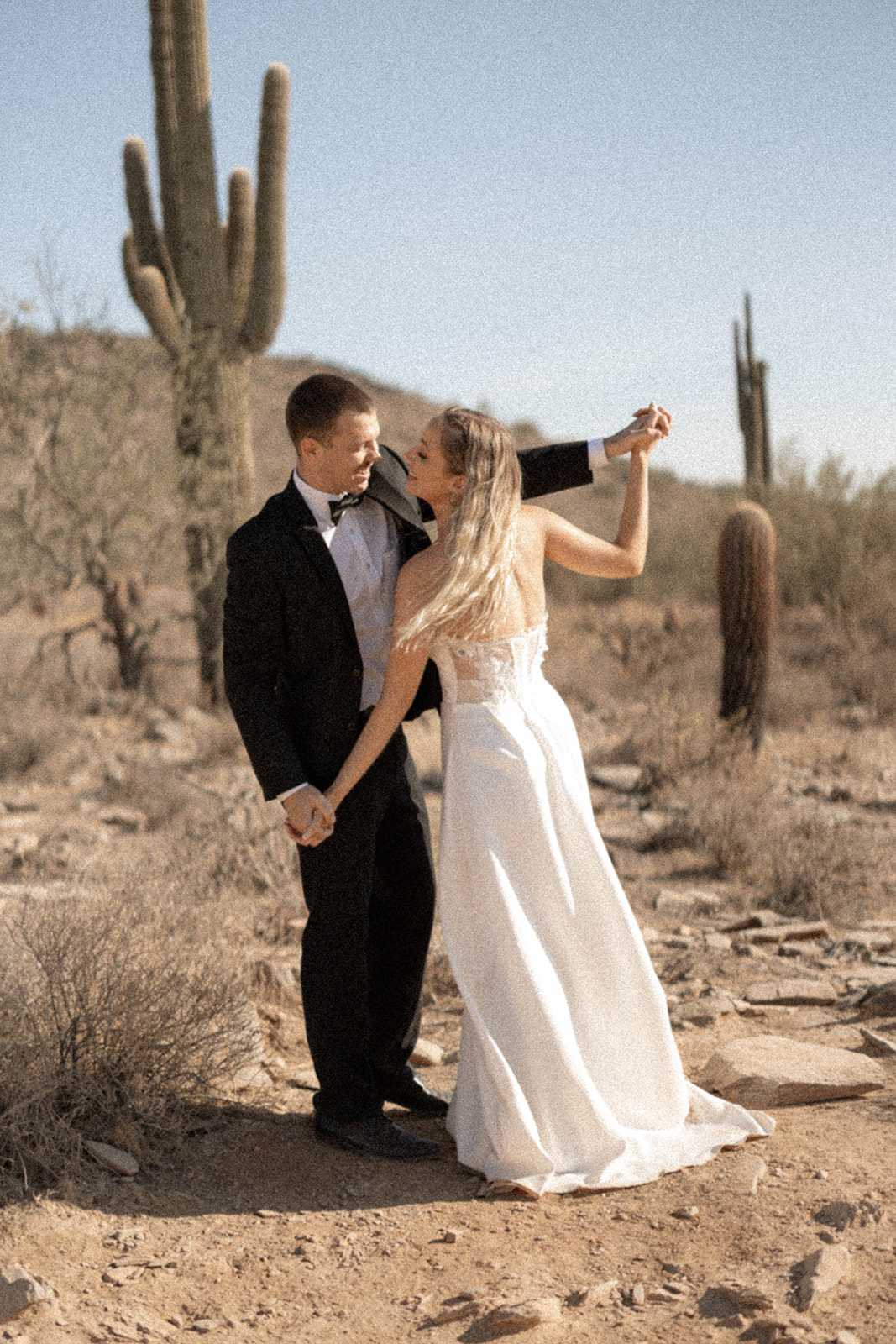 Bride and groom dancing in the desert with saguaro cacti in the background. The couple shares a playful and romantic moment as they hold hands and sway together, with the bride in a strapless white gown and the groom in a black tuxedo. The warm desert tones and soft sunlight create a relaxed and joyful atmosphere.
