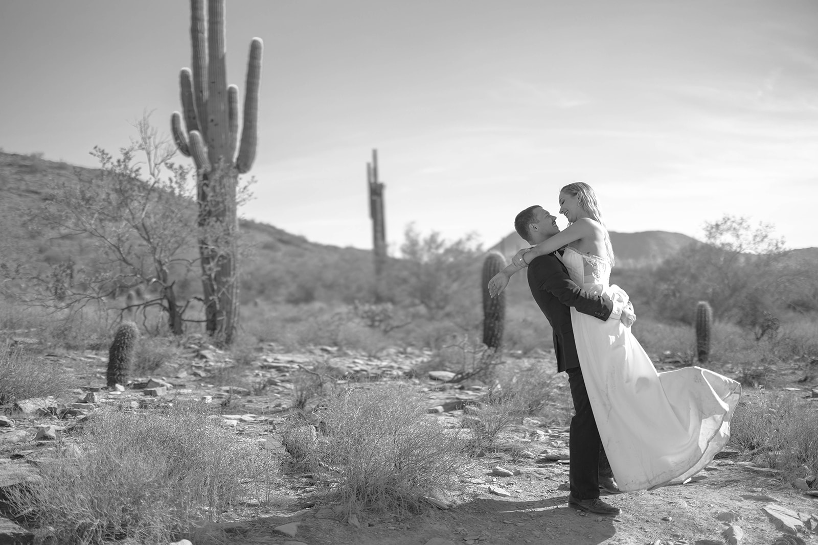 Groom lifting bride in the Arizona desert, surrounded by saguaros and rocky terrain.
