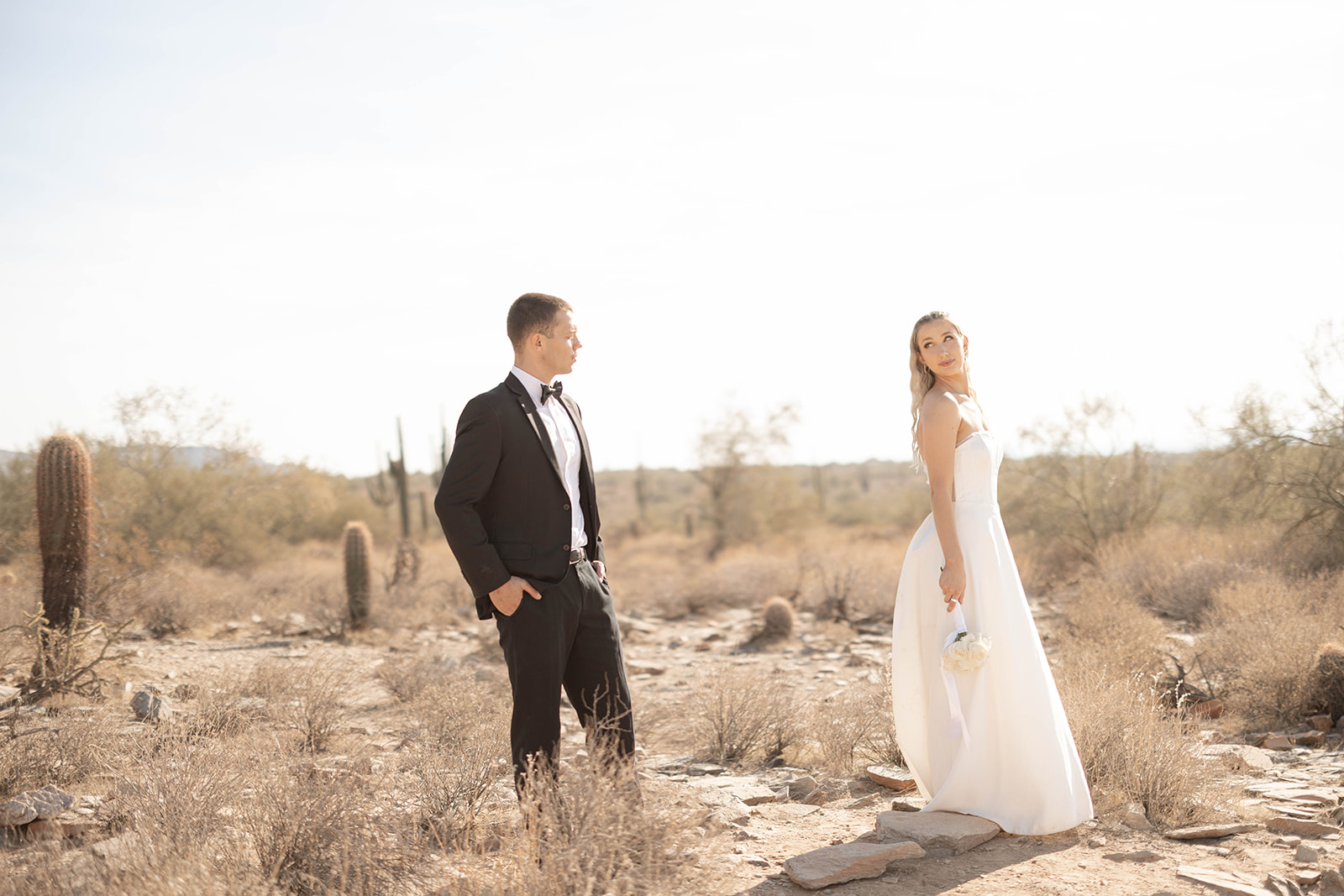 Bride glancing back at the groom while holding a small bouquet, walking through the serene desert landscape.