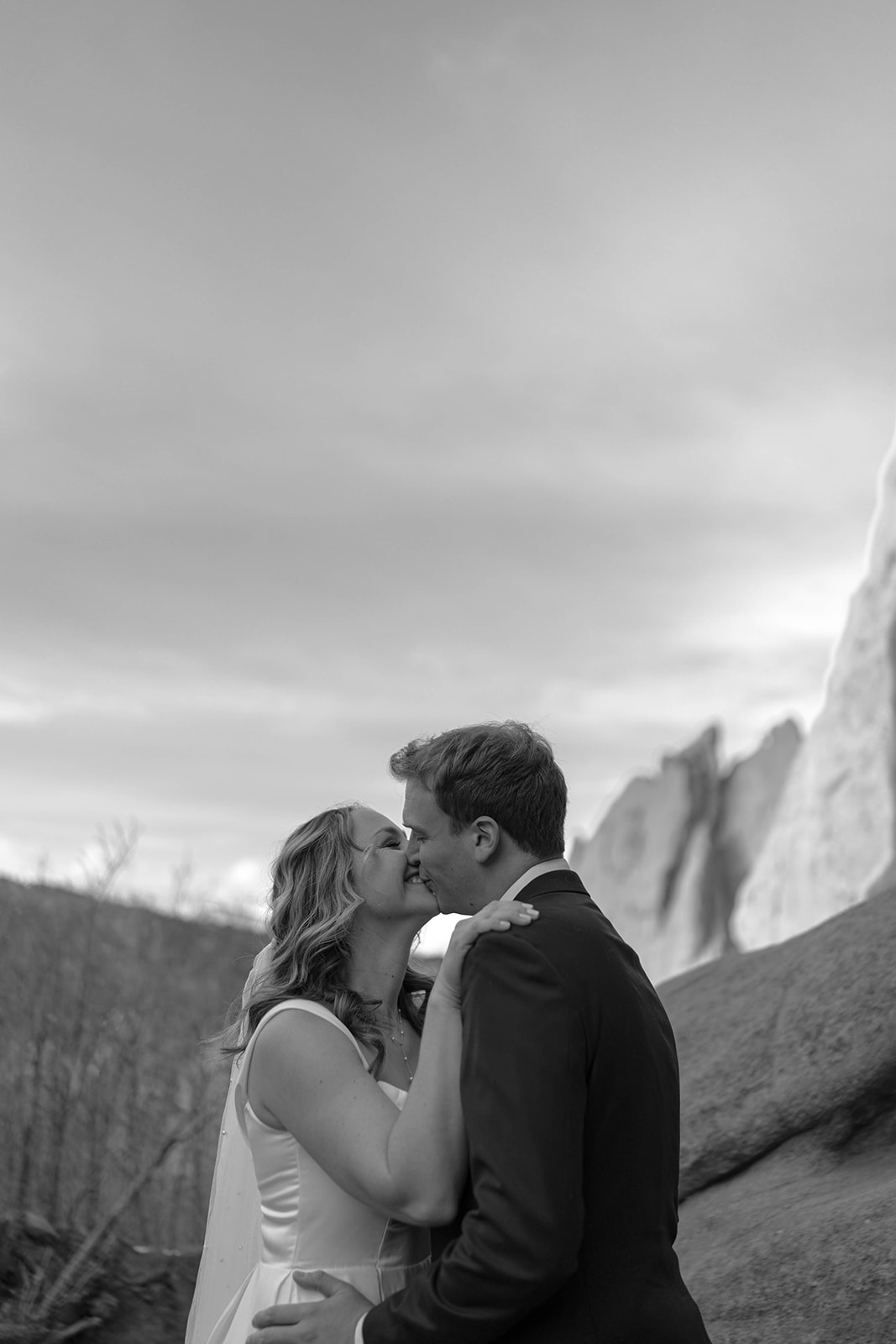 A soft black and white moment of a bride and groom leaning into each other with rocky cliffs rising behind them.