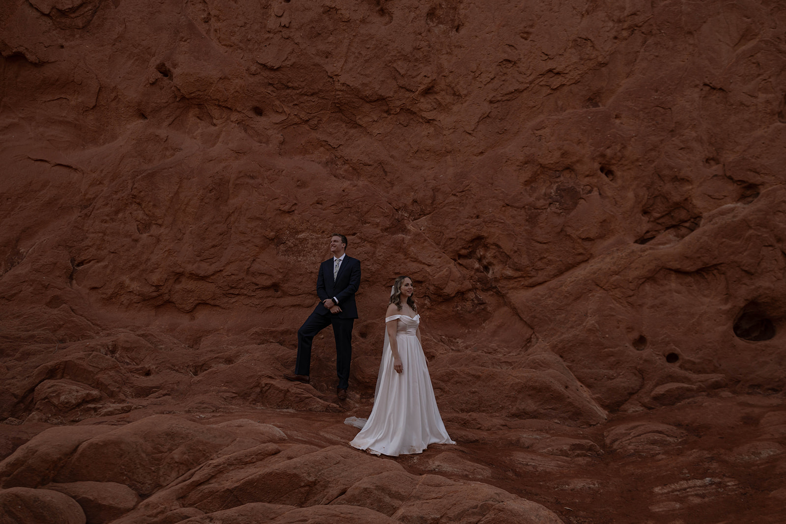 Couple standing against dramatic red rock cliffs, bride in off-shoulder gown.