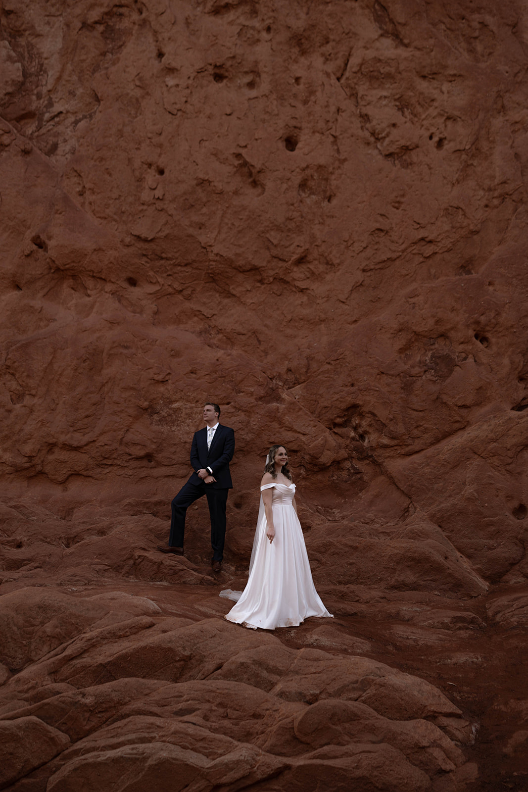 Bride and groom pose separately against a towering red rock formation, creating a bold and moody elopement portrait.