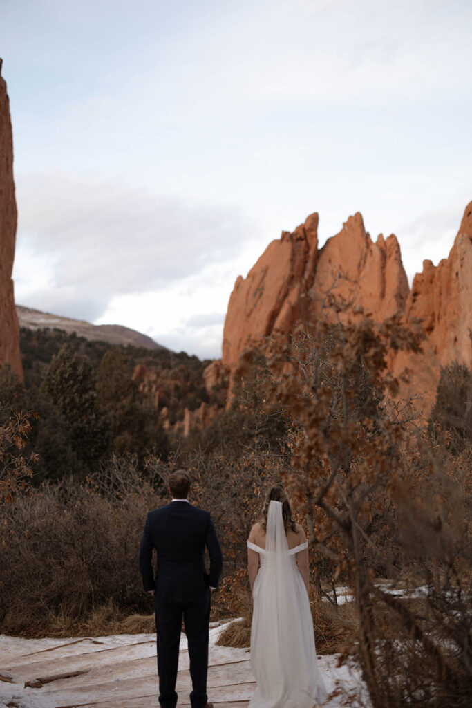 A couple stands facing towering red rock formations, their backs to the camera, taking in the dramatic landscape of one of the most iconic Colorado elopement locations.