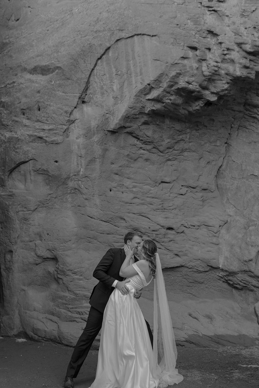 A black and white image of a bride and groom sharing a kiss in front of a textured rock wall, adding depth and intimacy to their elopement story.