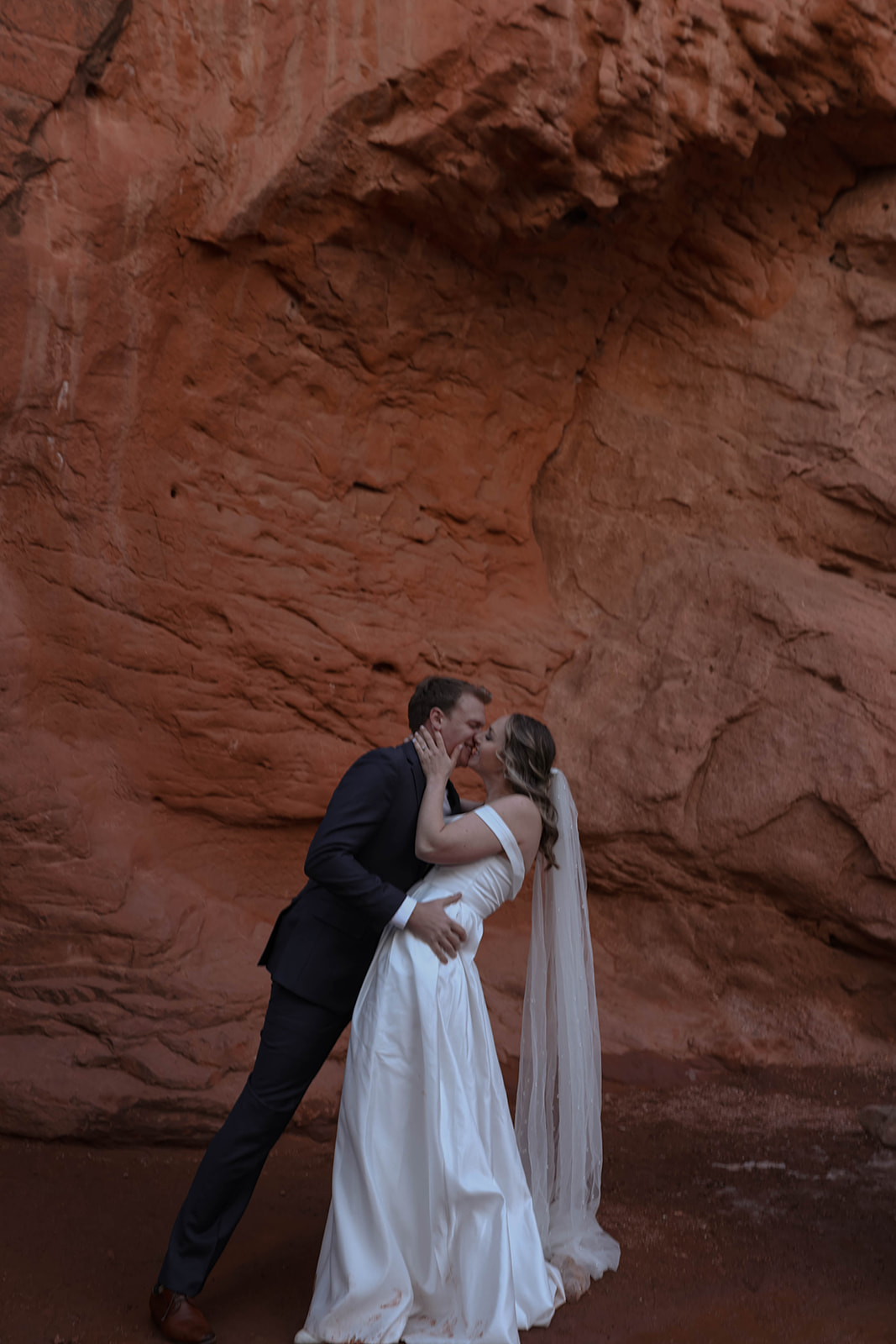 A couple kisses in front of a red rock wall, the bride’s veil flowing behind her as they celebrate their elopement in one of the dreamiest Colorado elopement locations.