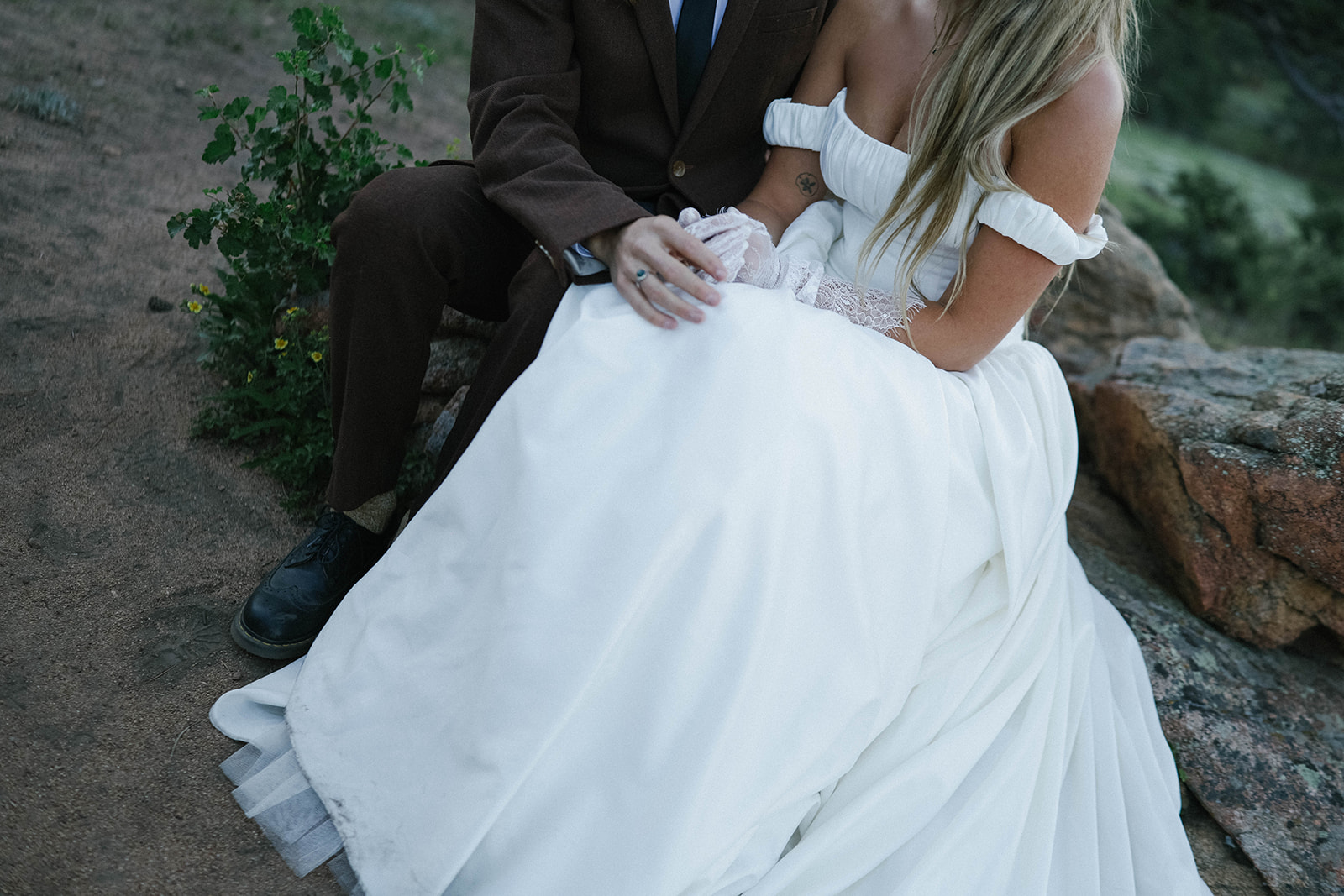 A seated bride and groom hold hands, her dress cascading around them while they sit on stone steps in the forest.
