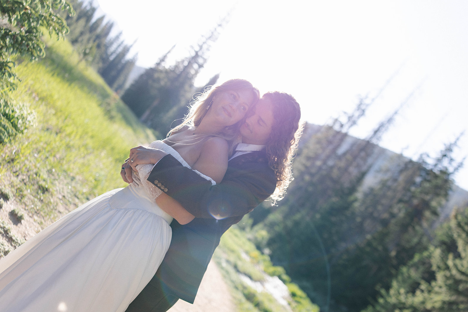 The couple embraces in glowing sunlight, surrounded by trees and a lush green field—an intimate moment in nature.