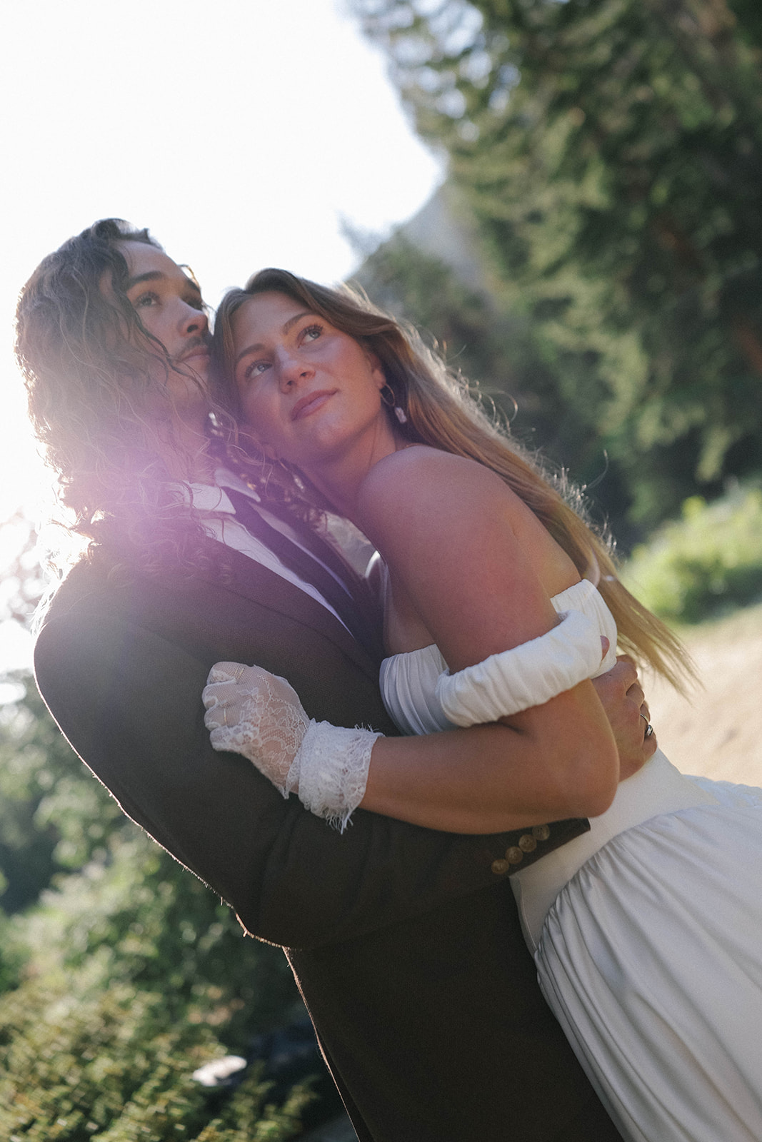 A bride and groom embrace in golden sunlight, their expressions soft and content as they take in the quiet beauty of the forest around them.