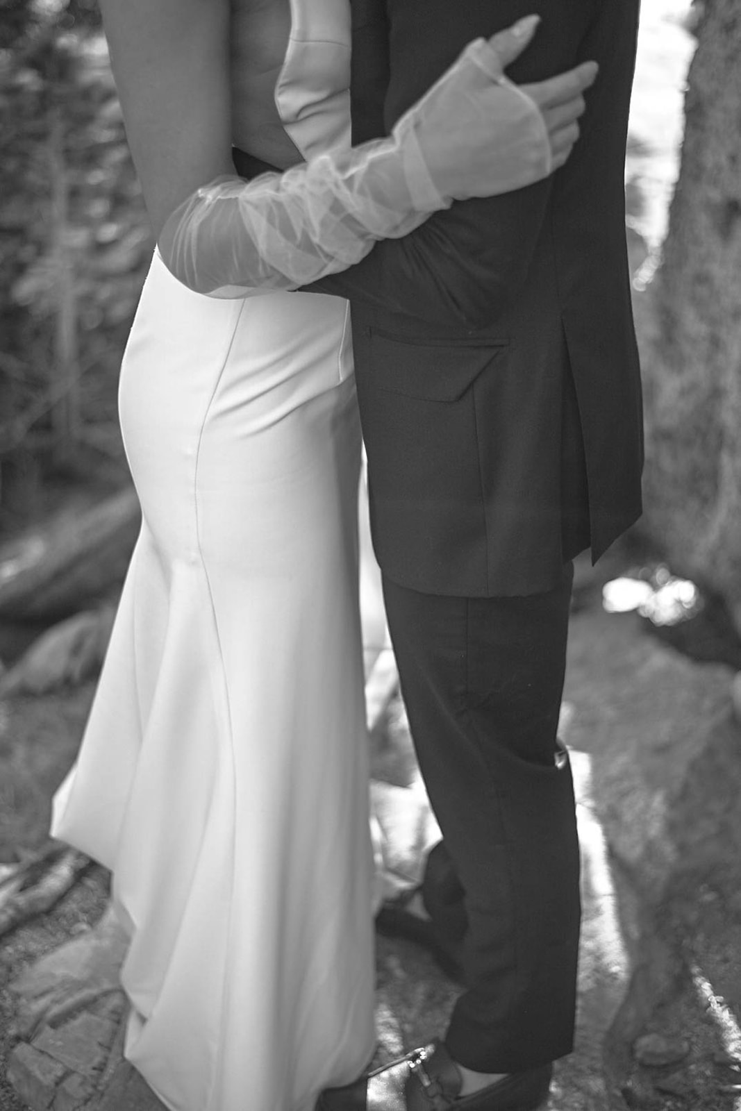 Black and white photo of a bride’s lace gloves wrapped around her groom during an intimate wedding moment.
