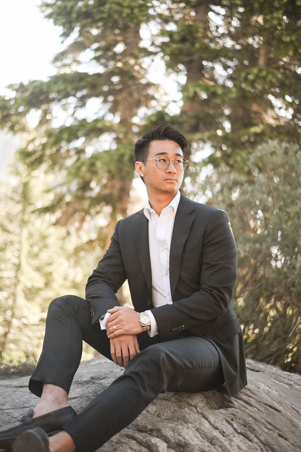 Groom in a black suit sits relaxed on a rock, surrounded by sunlit evergreens.