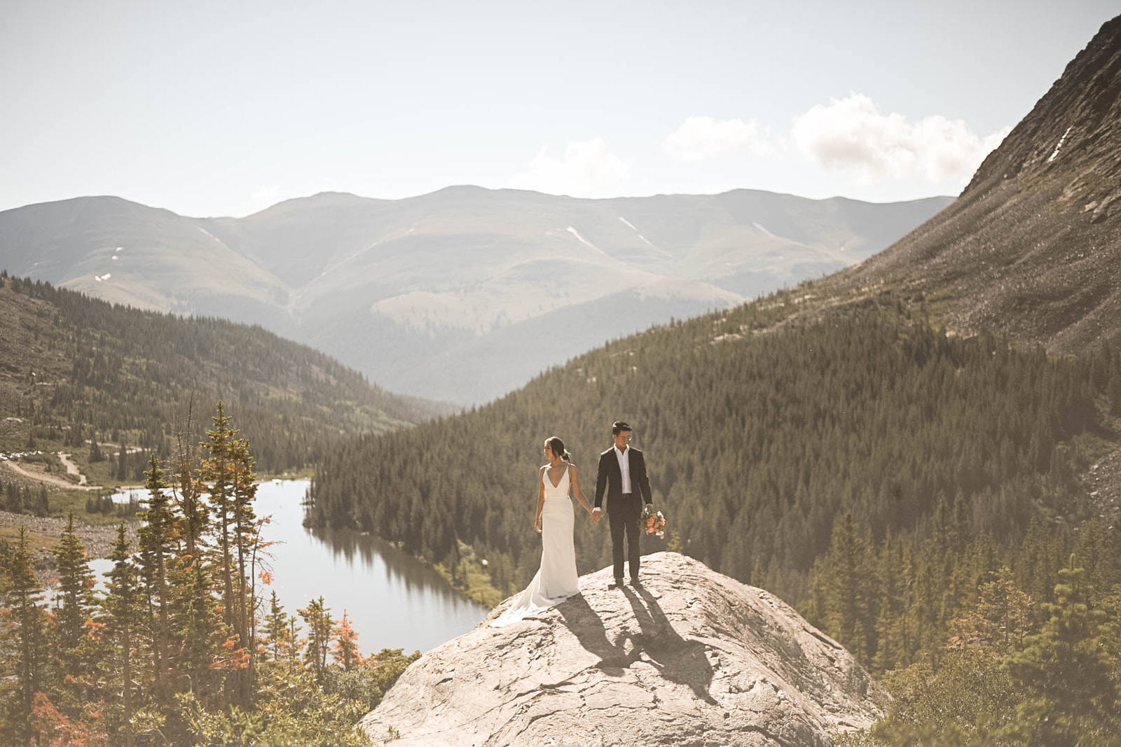 Wide shot of couple standing atop a mountain rock overlooking an alpine lake and forest, showcasing why summer mornings are the best time to elope in Colorado.