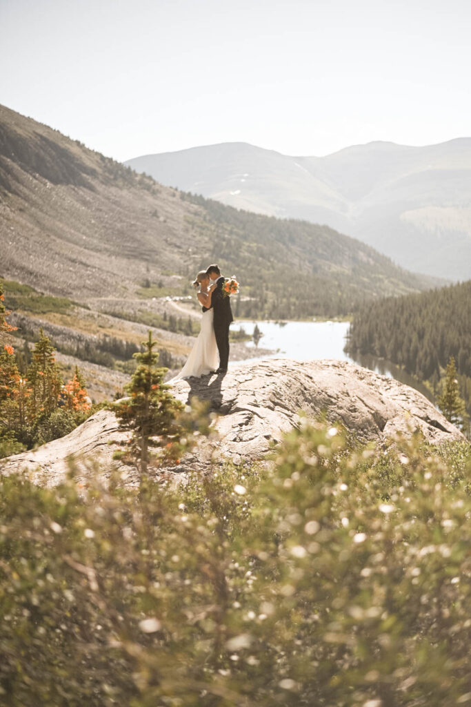 A couple embraces on a sun-drenched boulder overlooking a tranquil alpine lake and pine-covered valley, with mountain ridges stretching into the distance.