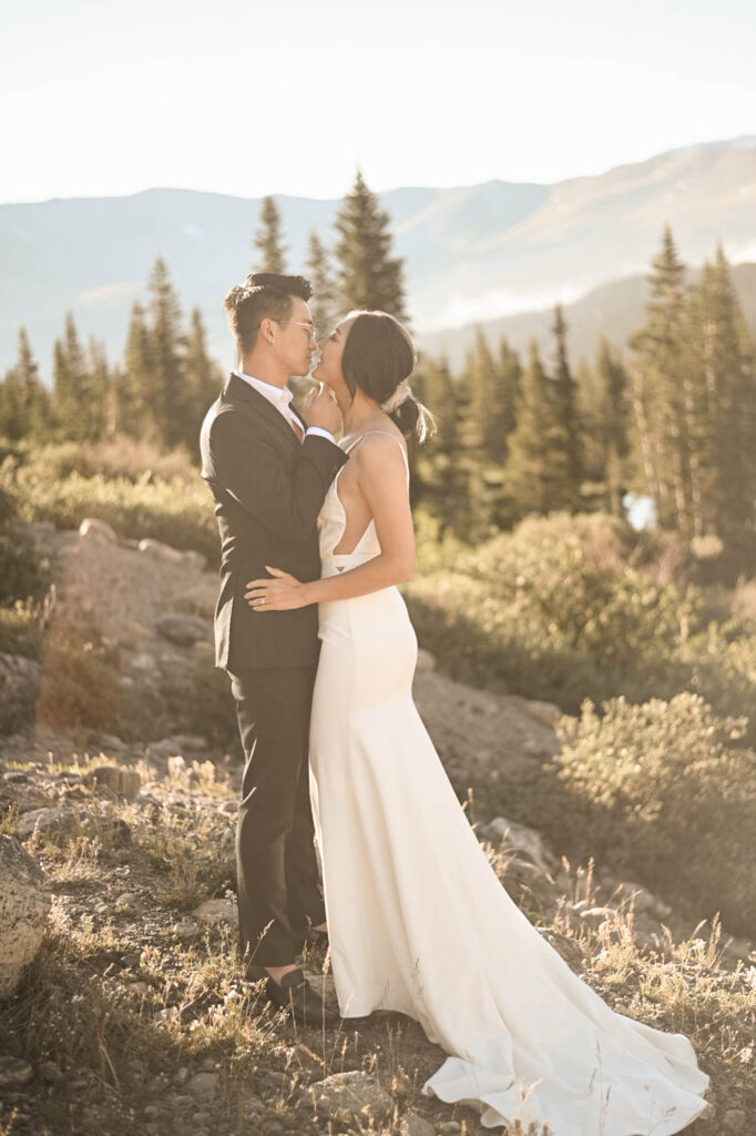 A bride and groom share an intimate moment on a sunlit mountain ridge, surrounded by evergreen trees and hazy alpine peaks in the distance.