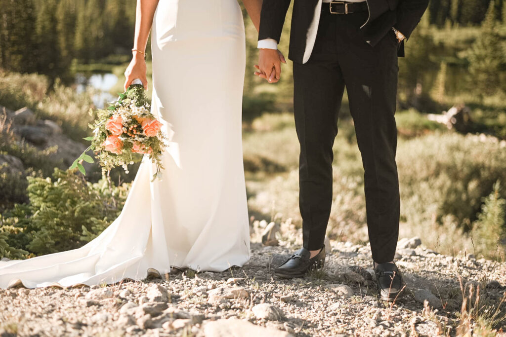 Close-up of a bride and groom holding hands in a forest clearing, highlighting the bride’s flowing ivory gown and bouquet of peach roses and greenery.
