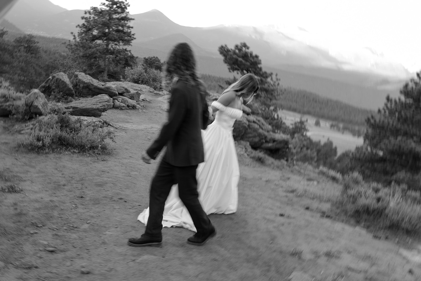 A black and white photo of a bride and groom walking hand in hand along a mountain trail, the bride slightly lifting her dress as they move together.