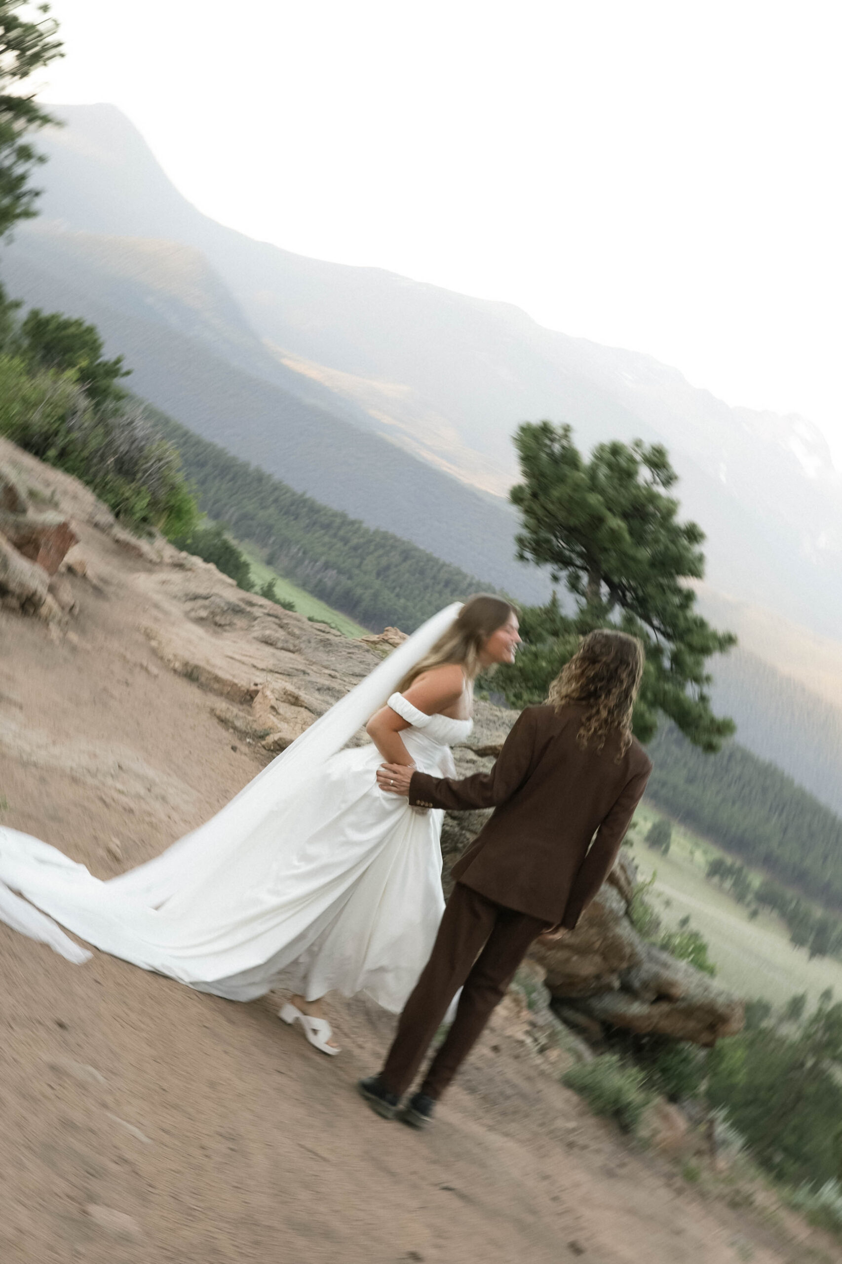The bride and groom walk hand in hand along a rugged path, her veil trailing behind, as the mountain landscape unfolds around them.