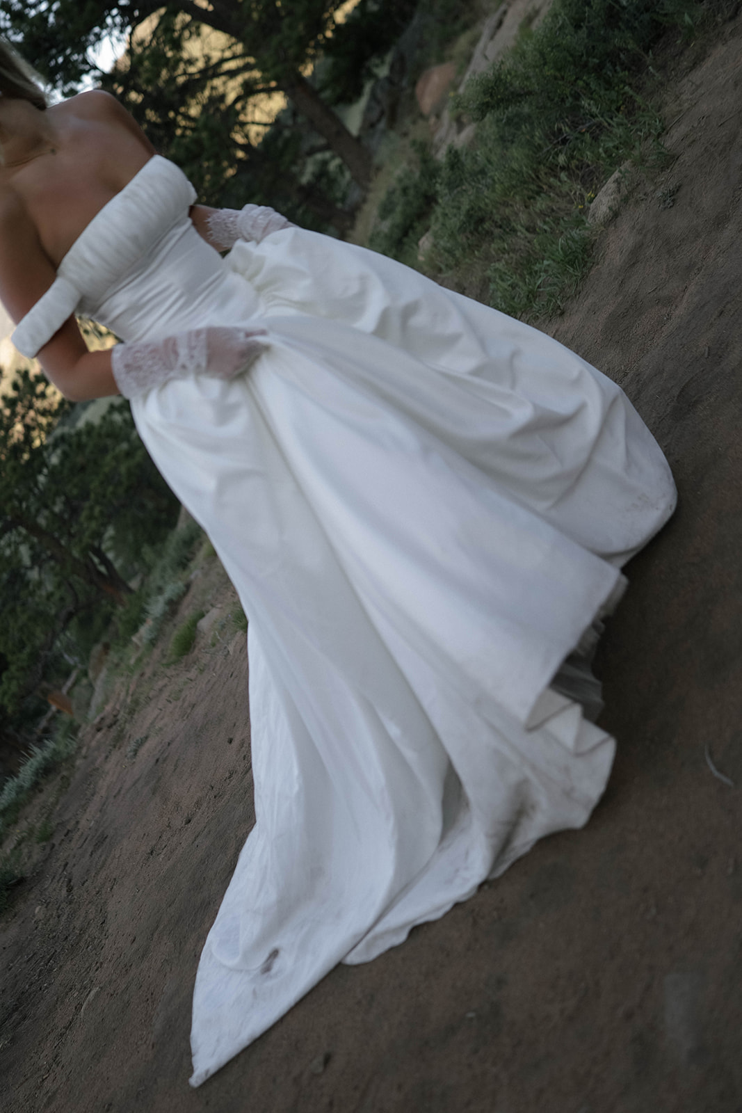 he bride lifts her dress slightly as she walks along a dirt trail through the forest, her gown flowing naturally with the movement.