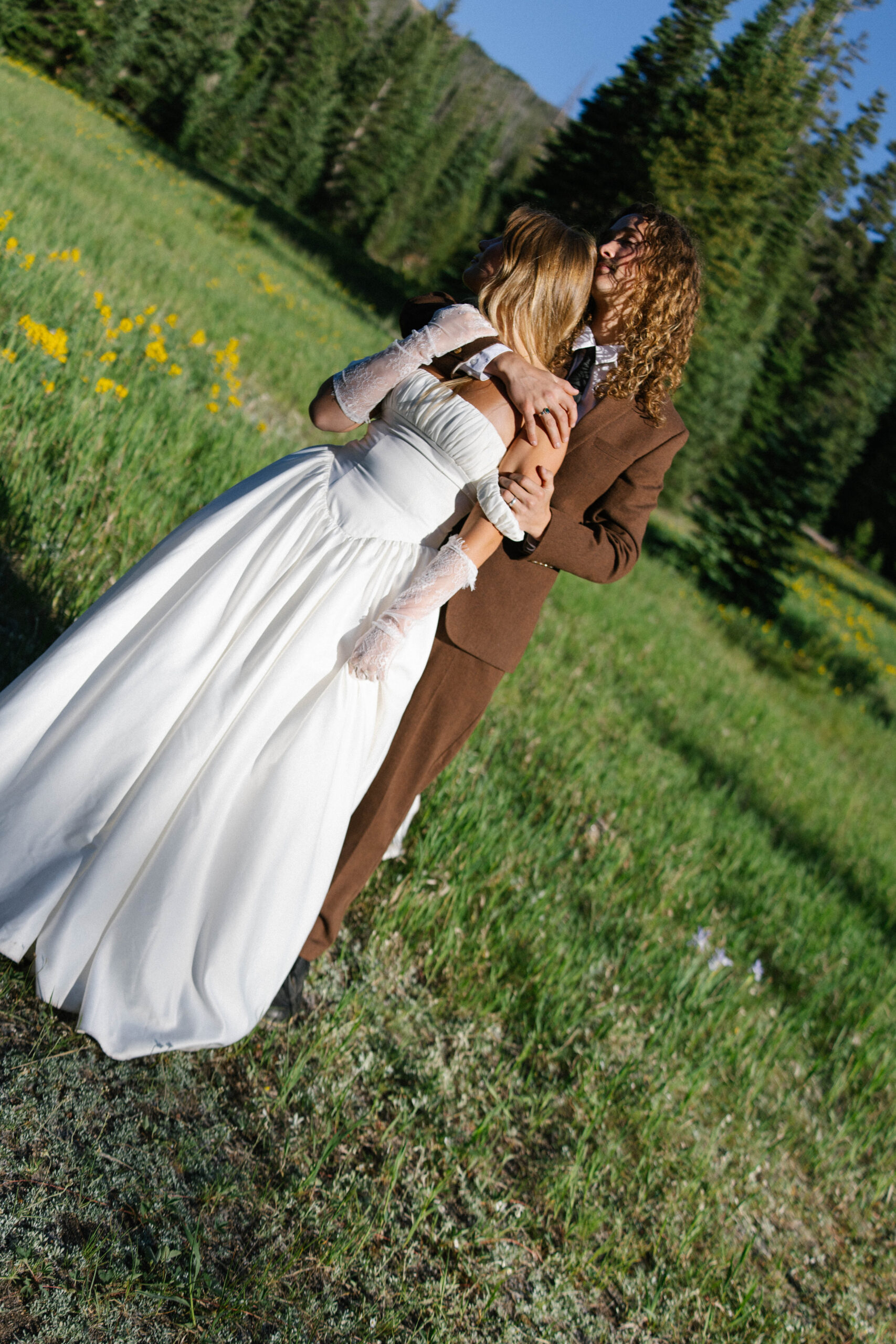 A couple embraces in a sunlit meadow surrounded by wildflowers and pine trees, sharing a tender moment during their outdoor elopement.