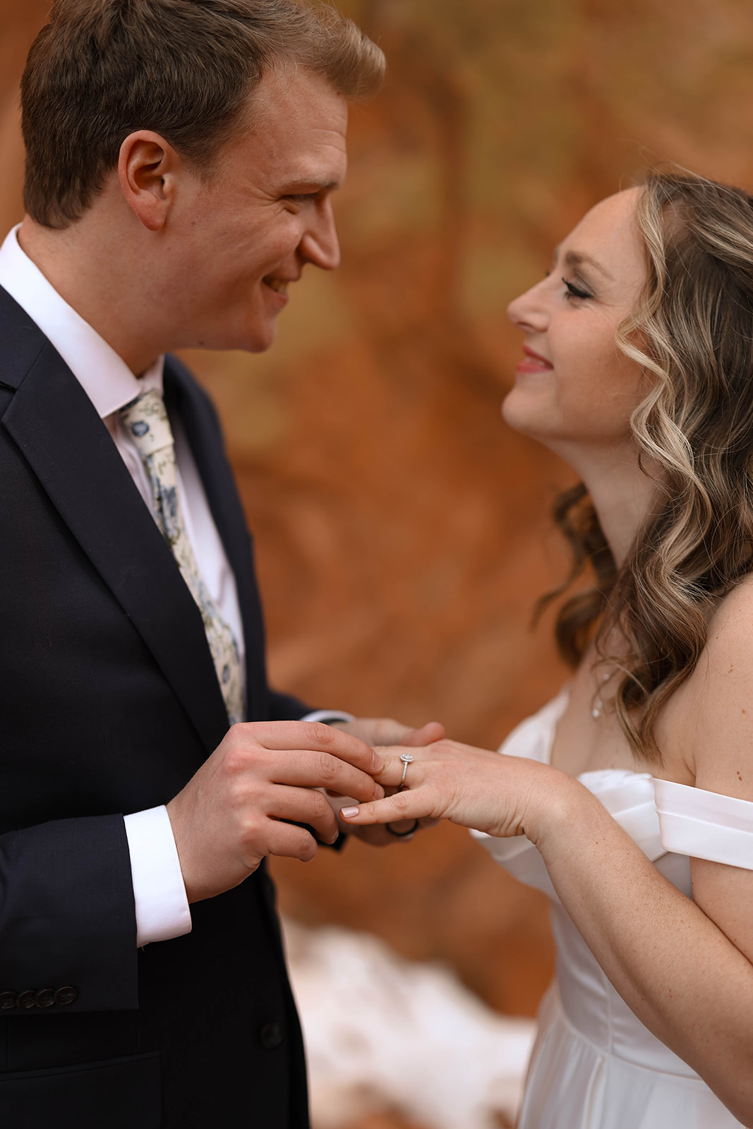 Groom places a wedding ring on the bride’s finger during their ceremony in front of warm red rocks, both smiling softly at each other—the best time to elope in Colorado is when the desert landscape glows like this.