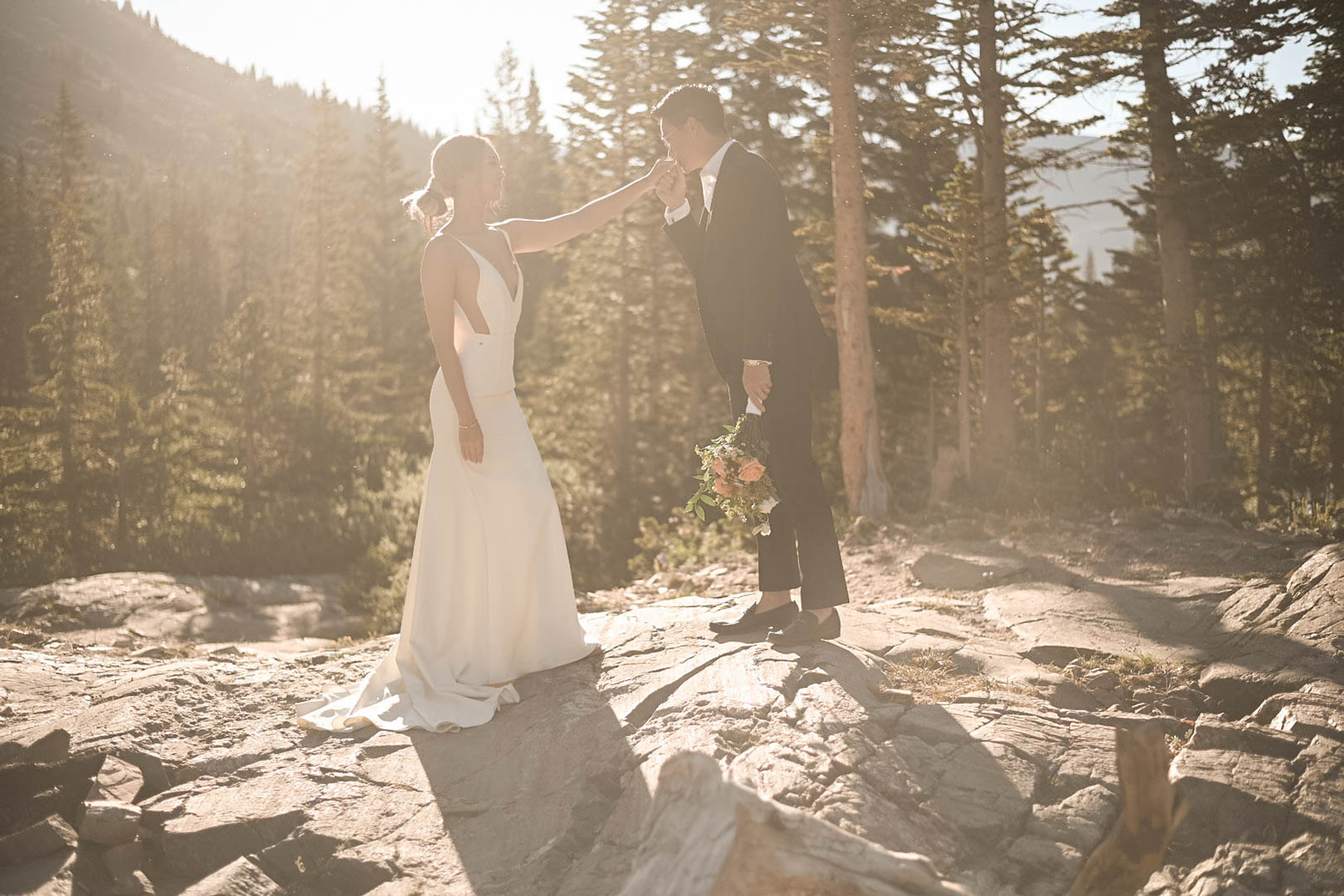 A groom gently kisses his bride’s hand on a sun-drenched rock outcrop, surrounded by evergreens—proof that summer might just be the best time to elope in Colorado.