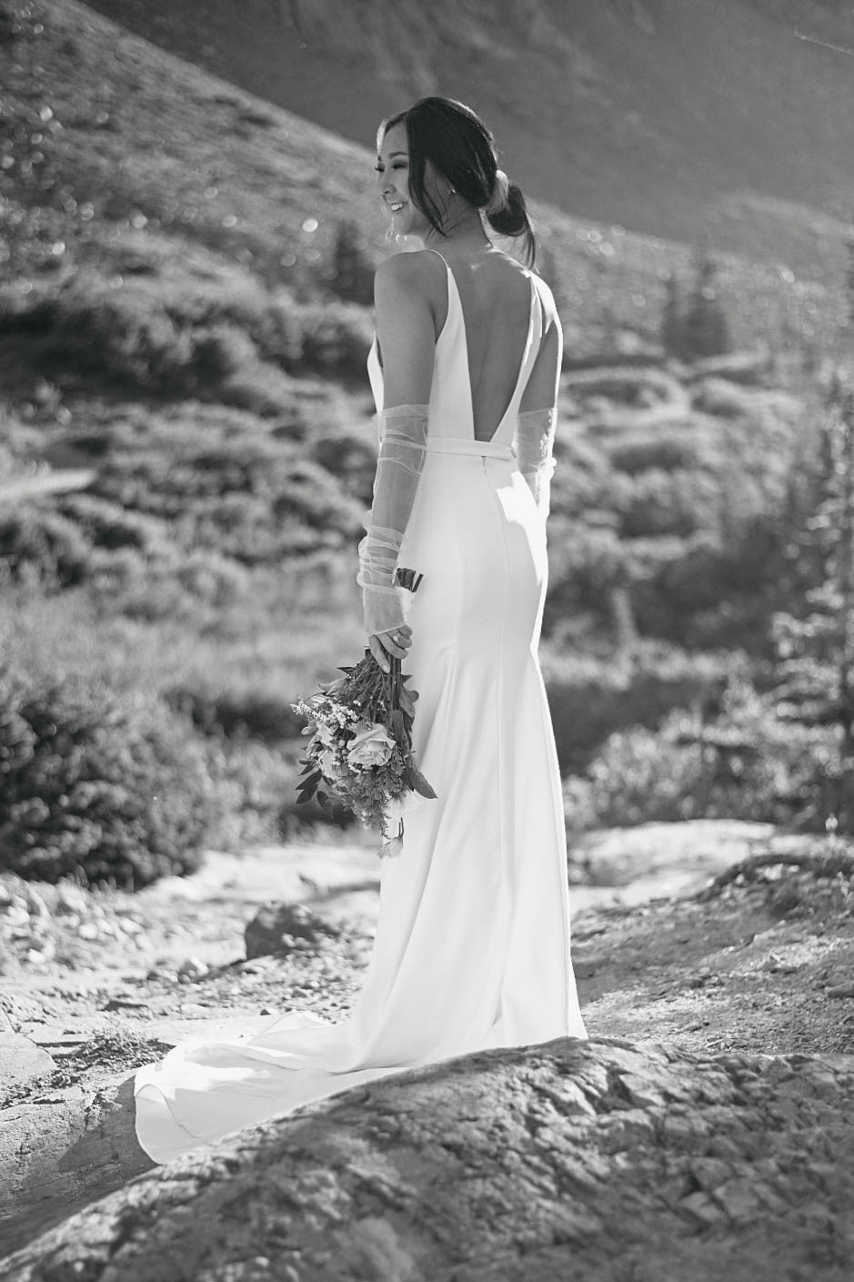 Bride smiling over her shoulder while holding her bouquet, standing on a rocky path—summer mornings like this are often the best time to elope in Colorado.