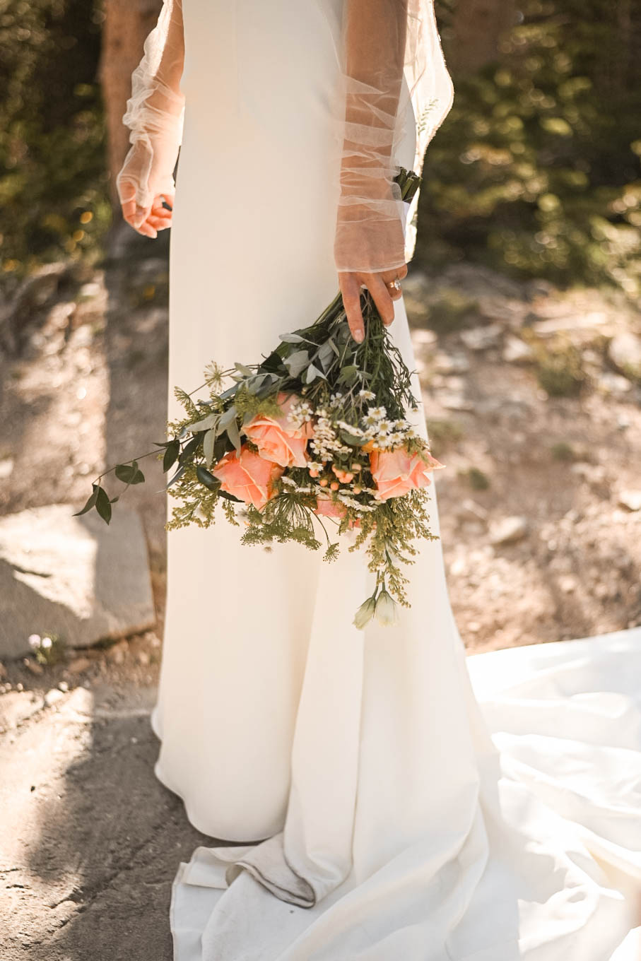 Close-up of a bride’s hand holding a peach rose bouquet, her satin gown trailing behind.