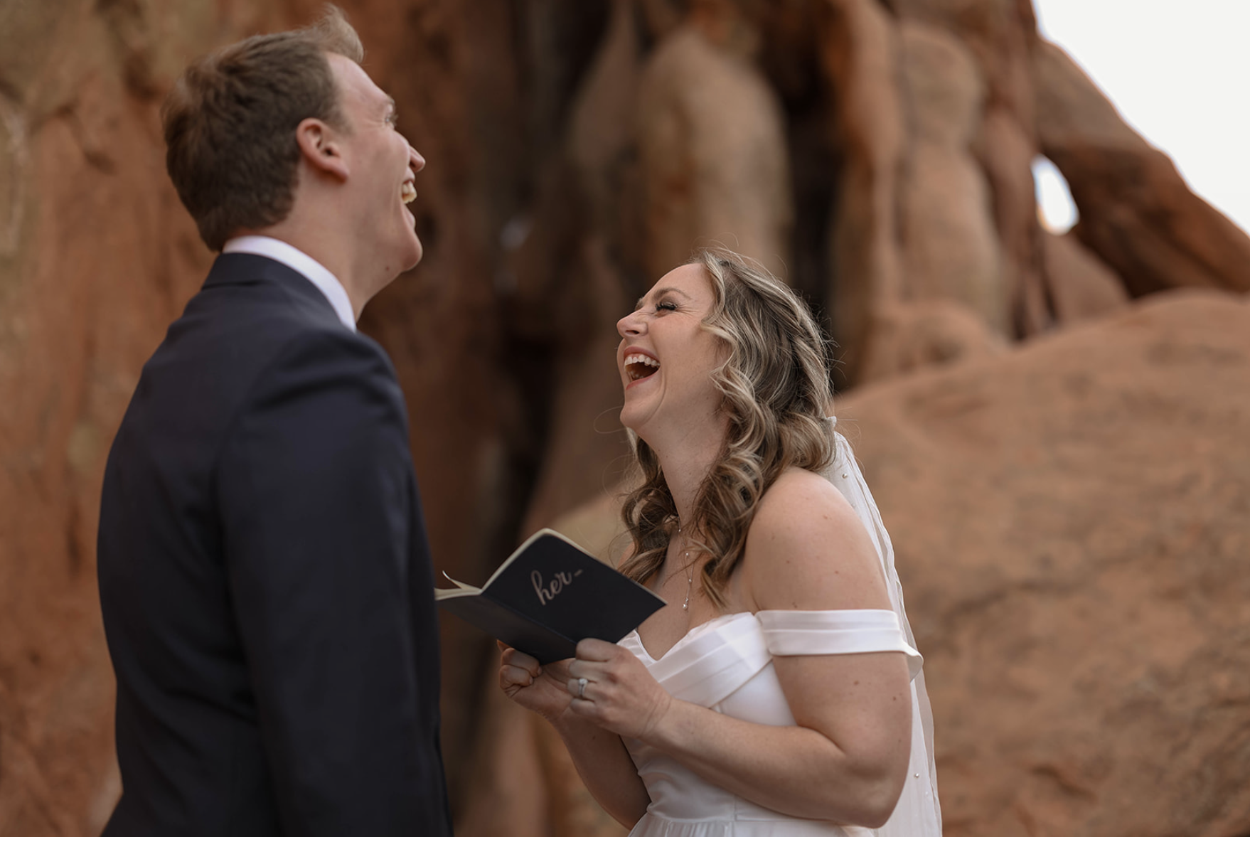 In a canyon of red rock, a bride laughs while reading her vows, a genuine moment that reflects the joy of choosing the best time to elope in Colorado.