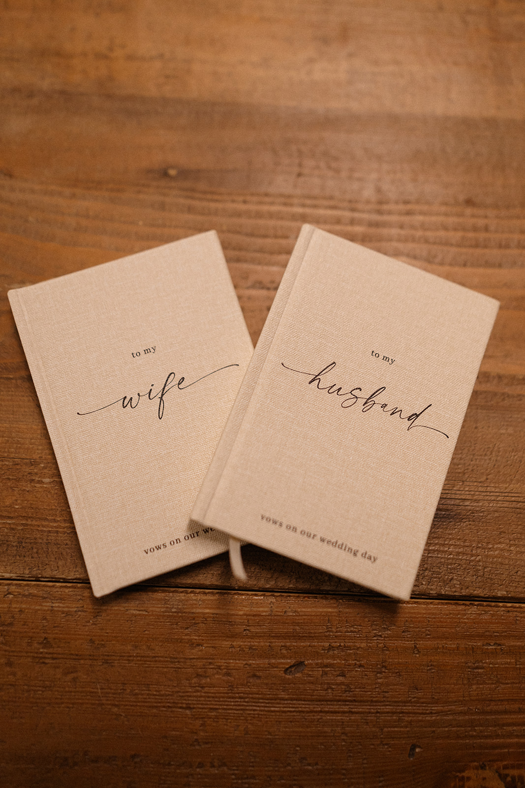 Beige linen vow books reading “to my wife” and “to my husband” rest on a rustic wooden table.