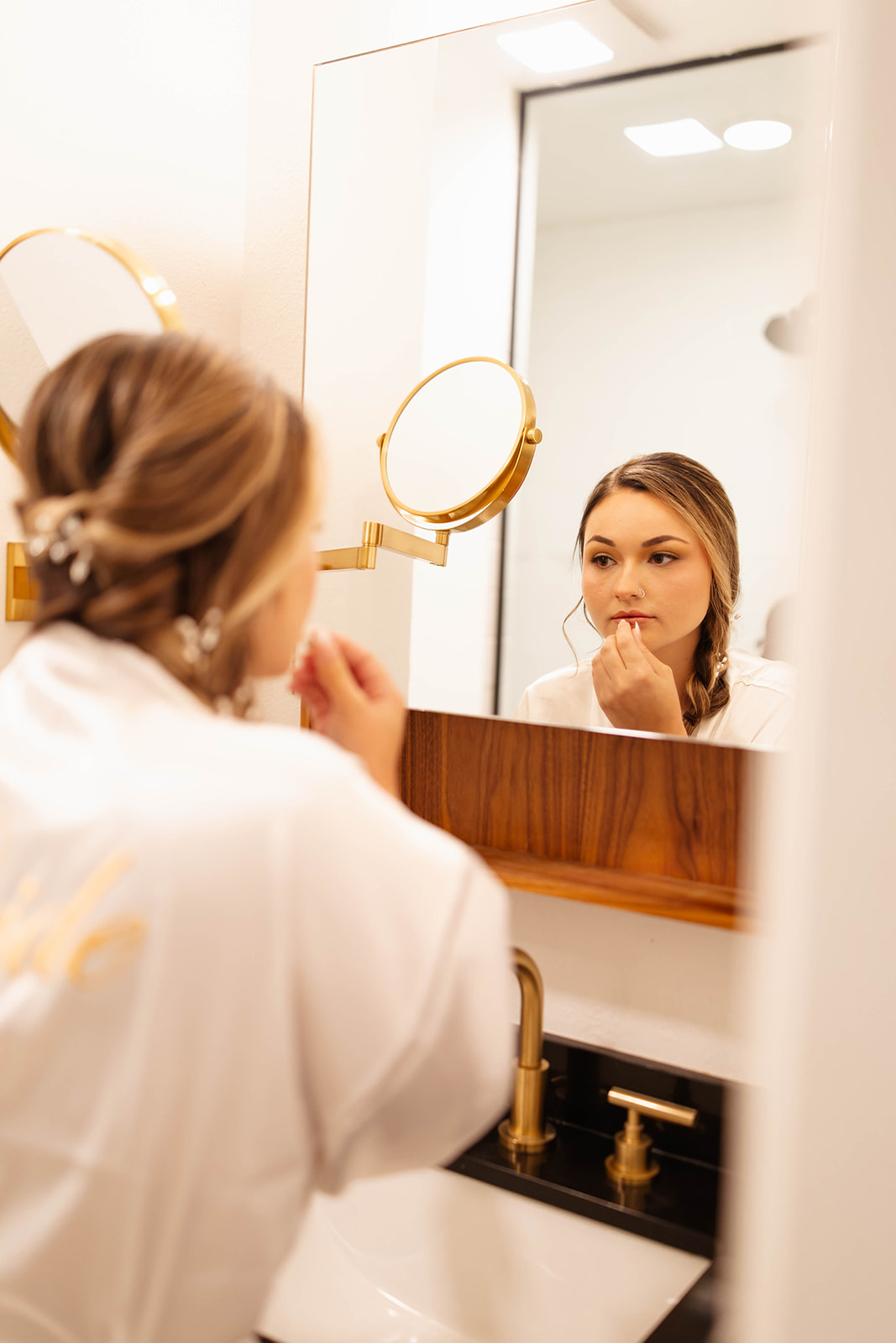 Bride in a silk robe applies lipstick while looking into a gold-framed mirror before the ceremony.